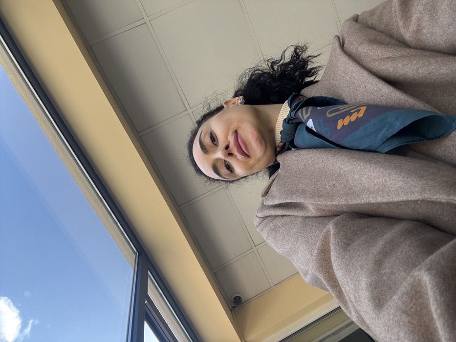 An adult female is depicted in a low-angle shot, looking directly up at the camera with a subtle smile. She has dark, curly hair and is wearing a light beige or camel-colored long coat over a cream or light yellow turtleneck. A dark teal silk scarf, patterned with abstract shapes in orange, white, and dark blue, is tied around her neck. A silver stud earring is visible in her right ear.
The background consists of a modern building facade featuring light beige paneling and a darker, horizontal upper band. To the upper left, a black-framed glass roof or canopy structure is visible. A small, white, spherical security camera is mounted on the building facade near the glass structure. Above the architecture, a clear blue sky with sparse white clouds indicates an outdoor setting. The environment is consistent with an urban area in the city of Tbilisi, Georgia, likely near a contemporary commercial or public building.