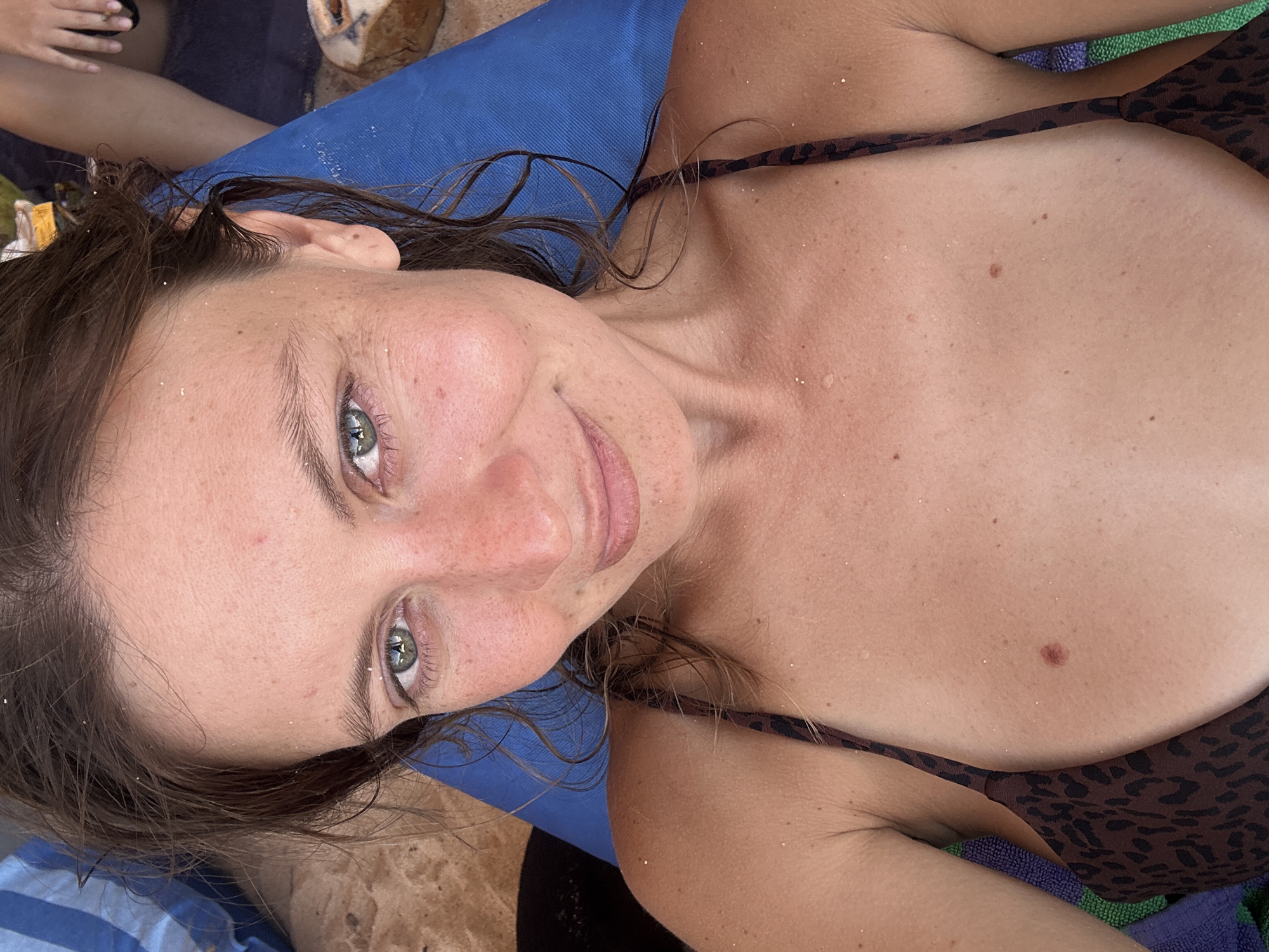 A woman with light skin and green eyes lies on a blue surface, seemingly a beach towel, with sand visible in her hair and on her skin. She wears a brown and black patterned bikini top. A coconut is visible in the background. The location is indicated as Kuta Selatan, Indonesia, suggesting a beach setting.