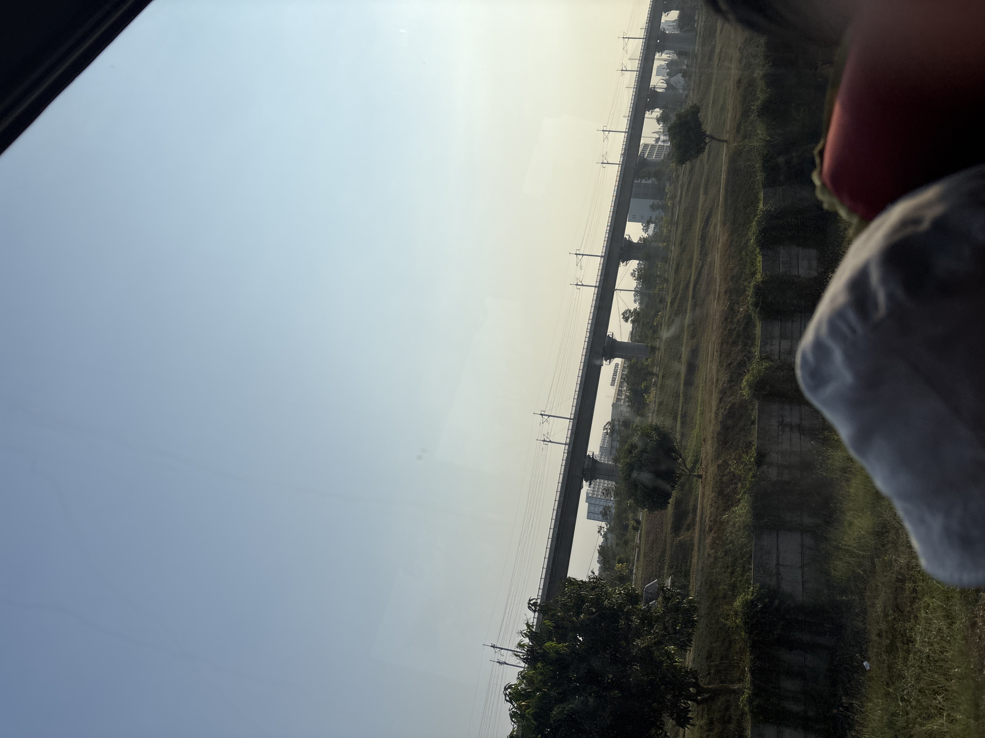 The image shows a view from inside a vehicle, looking out at a landscape with a raised metro line. The sky is clear and pale blue, with a slight haze near the horizon. The metro line, supported by concrete pillars, stretches across the frame. Below the metro line, there is a grassy slope with some trees and buildings in the distance. In the foreground, out of focus, are parts of what appear to be people's heads or shoulders, covered by grey and red fabric, suggesting they are passengers in the vehicle. The overall scene conveys a sense of travel or observation from a moving vehicle.