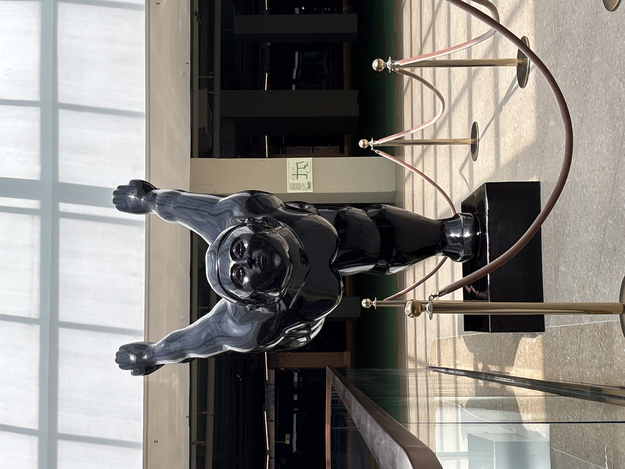 The image shows a large, black, polished sculpture of a person in a dynamic pose, with arms outstretched and raised. The sculpture is positioned in what appears to be a modern indoor space with a high ceiling and large windows that cast a grid of shadows on the floor. The sculpture stands on a black rectangular base, and is roped off with stanchions and dark red velvet ropes. In the background, there is a green floor, pillars, and some signage on a wall. The setting is an indoor public space, possibly a museum, gallery, or atrium. There is no indication that this location is in Saint Petersburg, Russia.