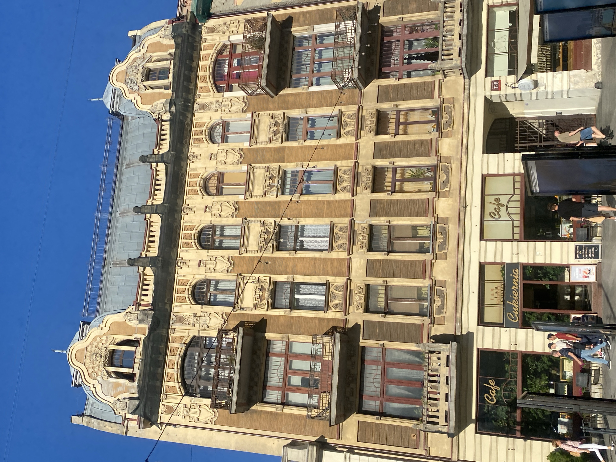 This is a vertical, eye-level, medium shot of an ornate, old building facade under a clear blue sky. The building, located in Łódź, Poland, features intricate architectural details, including decorative carvings around arched windows, balconies with wrought-iron railings, and a distinctive mansard roof. The facade is a mix of yellow and brown tones.
Several businesses are visible at the street level. One prominent sign reads "Cafe" in elegant script. Another sign displays "Cukiernia," indicating a confectionery or pastry shop. Above the entrance to one of the establishments, a small sign with the number "128" is visible.
There are a few people visible on the sidewalk in front of the businesses. Two men and a woman are standing near the cafe. Another person is leaning against the building, and two more individuals are walking on the sidewalk further down. The lighting suggests it is daytime, likely afternoon, with the sun casting shadows. The overall atmosphere appears to be casual and urban. The building's detailed facade and the street-level businesses suggest a historic city center.