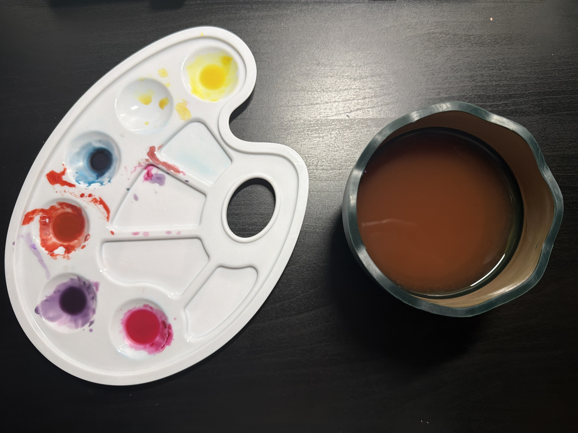 An overhead shot displays a white plastic paint palette and a container of discolored water set on a dark, textured surface. The kidney-shaped palette contains several distinct wells, holding vibrant paints including bright yellow, dark blue, bright red, deep purple, and a vivid pink/magenta. Smudges and drips of these colors, along with mixed hues like light blue and reddish-orange, are scattered across the palette's surface, indicating recent use.
To the right, a dark green container with an uneven rim holds brownish-orange, murky water, typically used for rinsing paintbrushes. The container's interior is a lighter beige or tan. Light reflects faintly off the water's surface. The background is a uniformly dark, possibly black, surface featuring a subtle wood grain or textured pattern, with some reflected light revealing minor dust particles. The arrangement suggests an ongoing or recently concluded painting activity. No people or visible text are present.