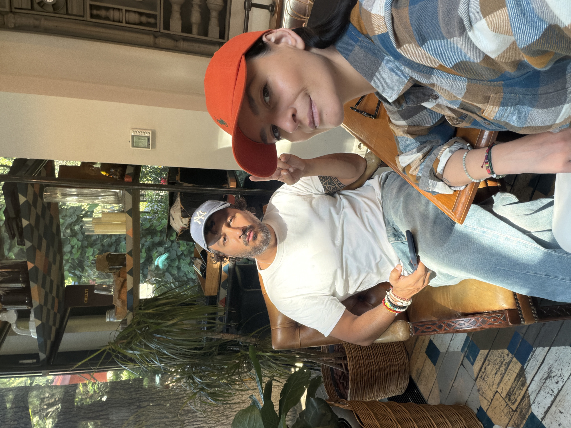 Two individuals, a male and a female, are captured in a selfie in Tbilisi, Georgia. The female is wearing an orange baseball cap and a plaid shirt. The male, seated in a leather chair, sports a white T-shirt, jeans, a baseball cap, and numerous bracelets. He is holding a phone. They are inside a cafe with a tiled floor, near a window with greenery outside, and a shelving unit with various items. The cafe has a rustic and somewhat vintage aesthetic.