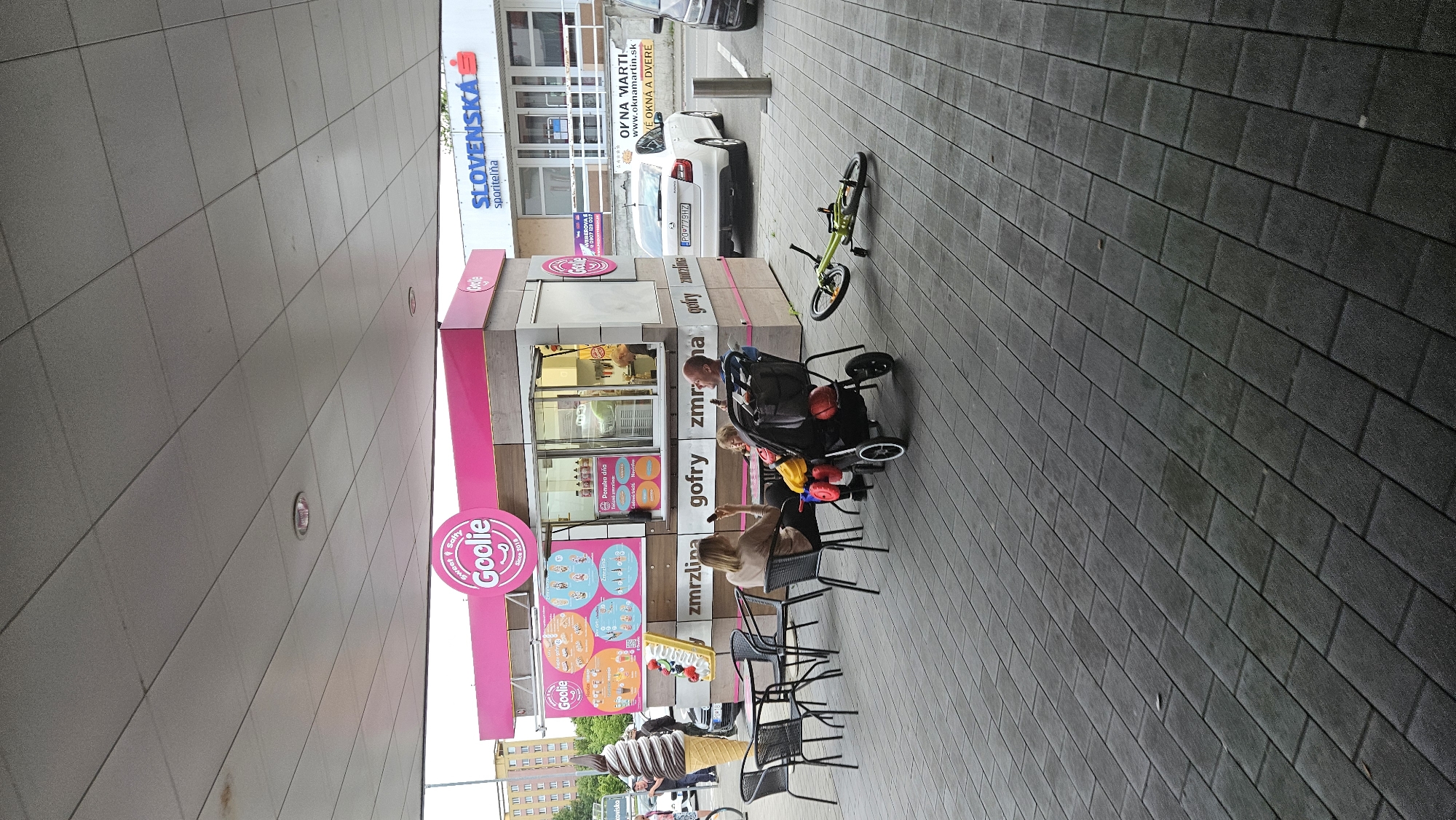 The image captures a casual street scene in Prešov, Slovakia, focusing on an outdoor food kiosk and its surroundings. The central feature is a vibrant pink and wood-paneled kiosk named "Goolie," which advertises "zmrzlina" (ice cream) and "gofry" (waffles) on its numerous signs and menu boards, some showing a "Ponuka dňa" (offer of the day). A large decorative ice cream cone stands prominently near the kiosk, reinforcing its specialty.
In the immediate foreground, several people are visible. Two women are seated at black metal tables, one engaged with her phone. A man stands beside a black baby stroller, which contains a child. A bright green BMX bicycle lies on its side on the grey paved ground. The entire area under the kiosk is covered by a large awning, suggesting a daytime setting with diffused light.
Beyond the kiosk, a commercial building displays the prominent blue sign of "SLOVENSKÁ sporiteľňa" (Slovak Savings Bank). Further down the street, a white car with the Slovak license plate "P00779HZ" is parked. Another sign for "OKNA MARTIN" (windows and doors), including its website "www.oknamartin.sk" and the phrase "VÉ OKNÁ A DVERE" (big windows and doors), along with a phone number "WEBEROVAS 0907 129 00," is visible on a building. The paved walkway extends into the background, where more buildings and vehicles can be seen, depicting a typical urban environment. The scene conveys a relaxed, everyday atmosphere.