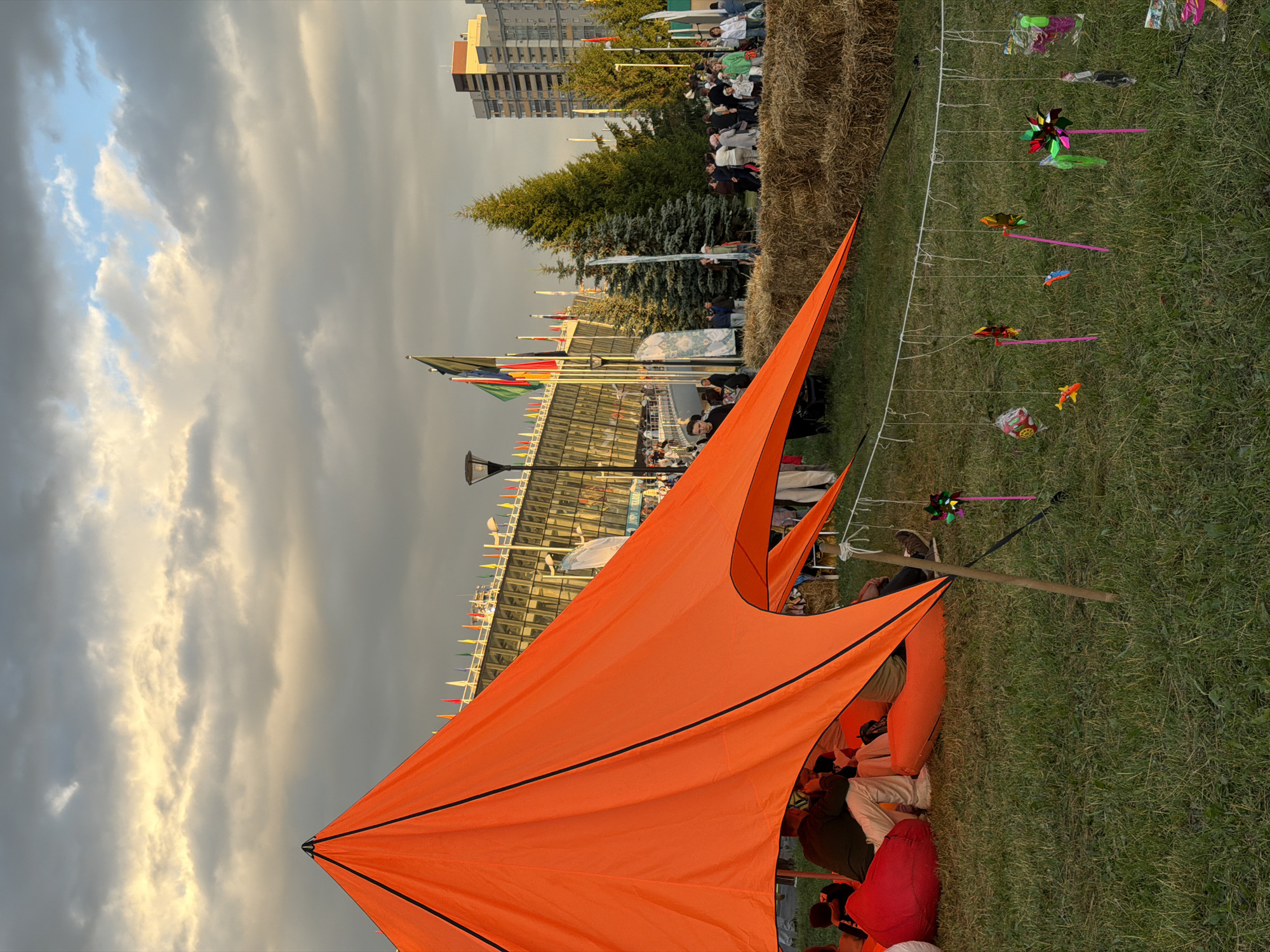 The image depicts an outdoor event with a large orange tent in the foreground. Several people are seated or reclining under the tent, some on beanbag chairs. The ground is covered in green grass. In the midground, there are bales of hay and a line of decorative items, including pinwheels and other small trinkets, hanging from a string. Beyond the hay bales, a crowd of people are gathered, with a modern building complex featuring glass facades and numerous flags visible in the background. Several trees, including evergreen species, are interspersed throughout the scene. In the far background, taller residential buildings can be seen. The sky is cloudy with patches of yellow light, suggesting either sunrise or sunset. The overall impression is of a festive gathering in an urban or semi-urban park setting.