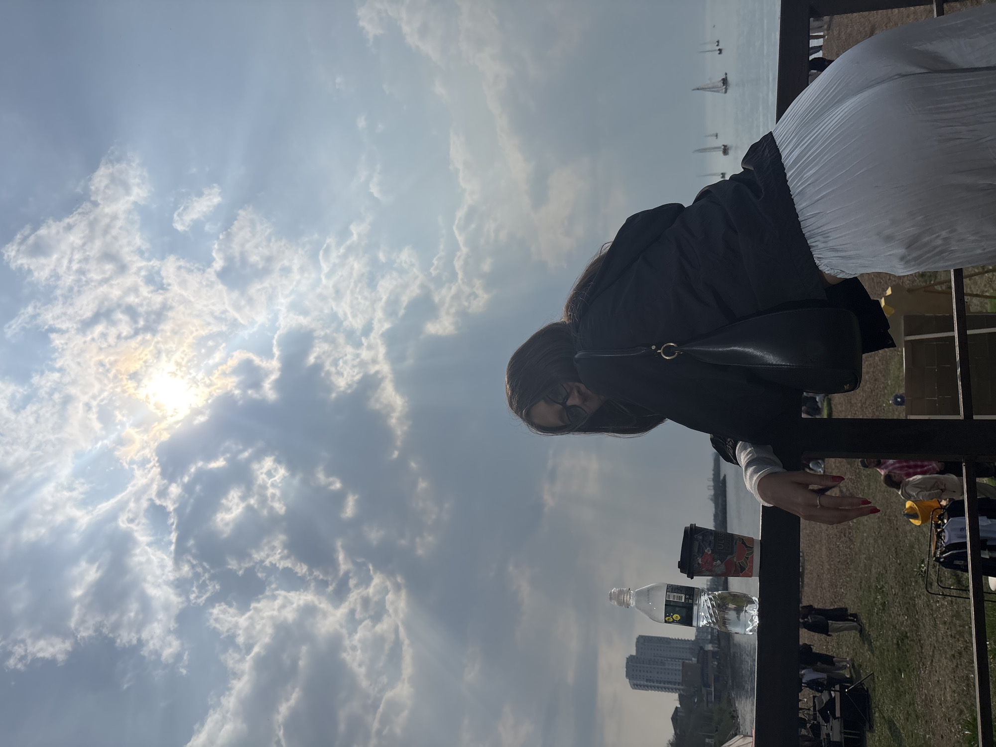 A woman stands facing a cityscape in Vladivostok, Russia, under a sky filled with clouds and sunbeams. She wears a black jacket, a white skirt, and sunglasses, with a black purse slung over her shoulder. She is leaning on a black railing next to a bottle of water and a cup. In the background, high-rise buildings are visible. Sailboats dot the sea in the distance. People are dispersed on the grassy area below.