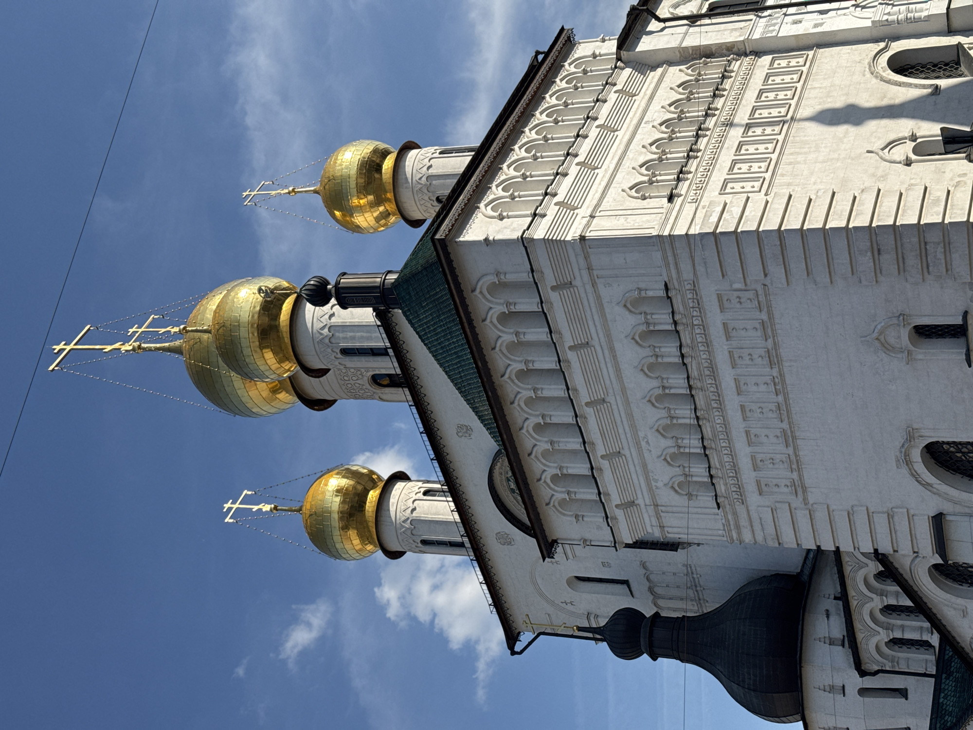 A low-angle, outdoor, medium shot captures the exterior of a white Orthodox church under a clear blue sky with scattered white clouds. The church features multiple golden domes topped with crosses, and a prominent black dome with a small golden cross. The white stone facade is adorned with intricate architectural details, including arched windows and decorative patterns. A shadow of the church's steeple is cast on the side of the building. The scene is devoid of people.