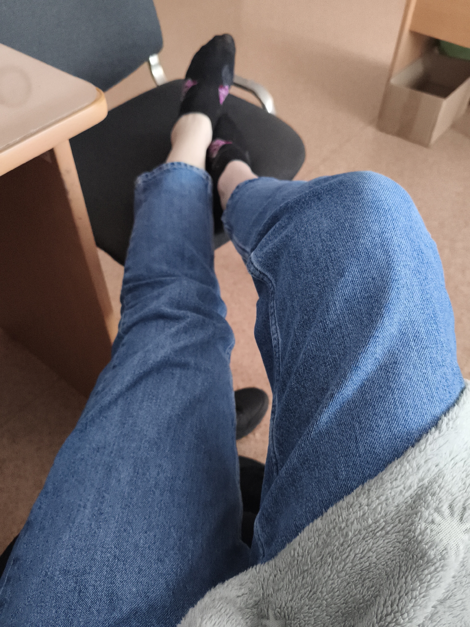 The image presents a view from the perspective of someone seated, displaying their legs crossed at the knees. The person is wearing blue jeans and black socks with pink patterns. Their feet are resting on a black office chair, which sits adjacent to a light brown desk. The background includes a cardboard box on the floor, and the presence of a grey, fleece-like material suggests the person is also wearing a sweater or has a blanket. The location is indoors, likely an office setting.