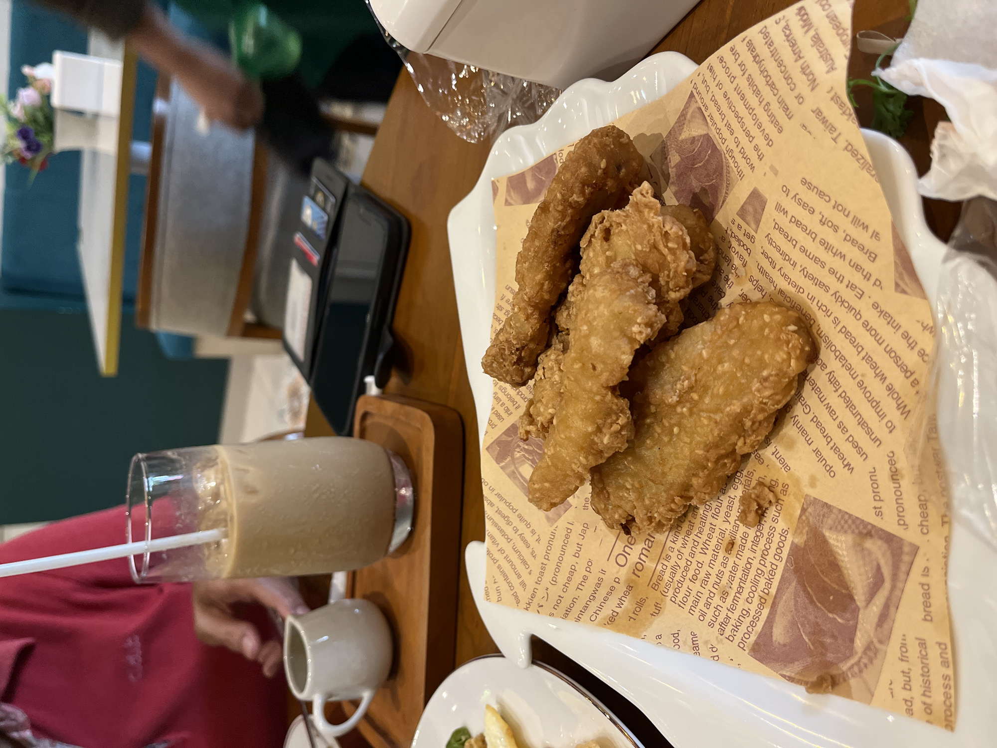 A close-up, eye-level shot shows a white plate with fried food, possibly chicken tenders, resting on a brown paper liner. The paper liner has text printed on it, appearing to be a historical or informational piece about bread. In the background, a tall glass filled with iced coffee is visible, along with a small white creamer pitcher on a wooden tray. A person's left arm and hand are partially visible on the left side of the frame, reaching towards the creamer. The setting appears to be a restaurant or cafe, with a blurred background showing tables and seating. The overall lighting is natural, with some overhead illumination.