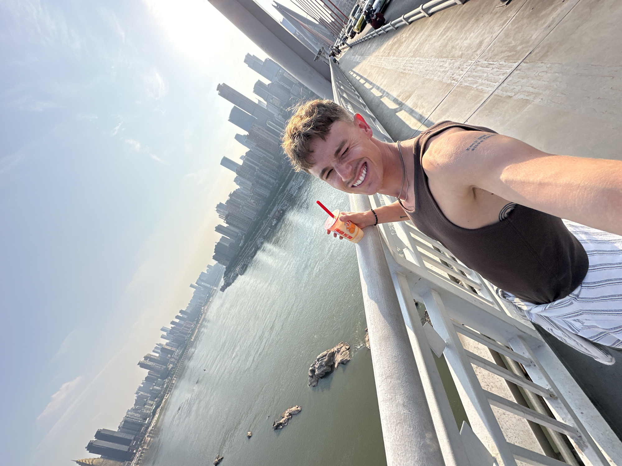 A man smiles while taking a selfie in Yuzhong District, China. He is on a bridge looking out over the water. He is leaning against the railing, holding a drink with a straw. He is wearing a tank top and striped pants. Behind him are the skyscrapers of the city.