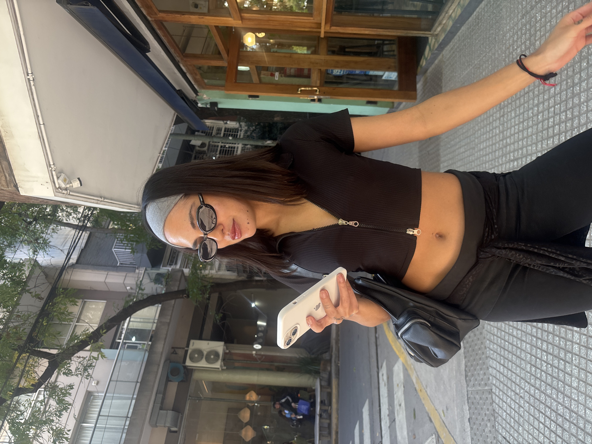A woman stands on a sidewalk in Buenos Aires, Argentina, holding a phone. She wears a black cropped top with a zipper, black pants, a gray headband, and sunglasses. She also has a black wristband and a small black bag. The sidewalk has a patterned surface. Behind her, there's a building with multiple windows, air conditioning units, and a tree. Across the street, there's a cafe or restaurant with a wooden frame. The sky is bright. The woman is holding the phone in her left hand and her right hand is raised.