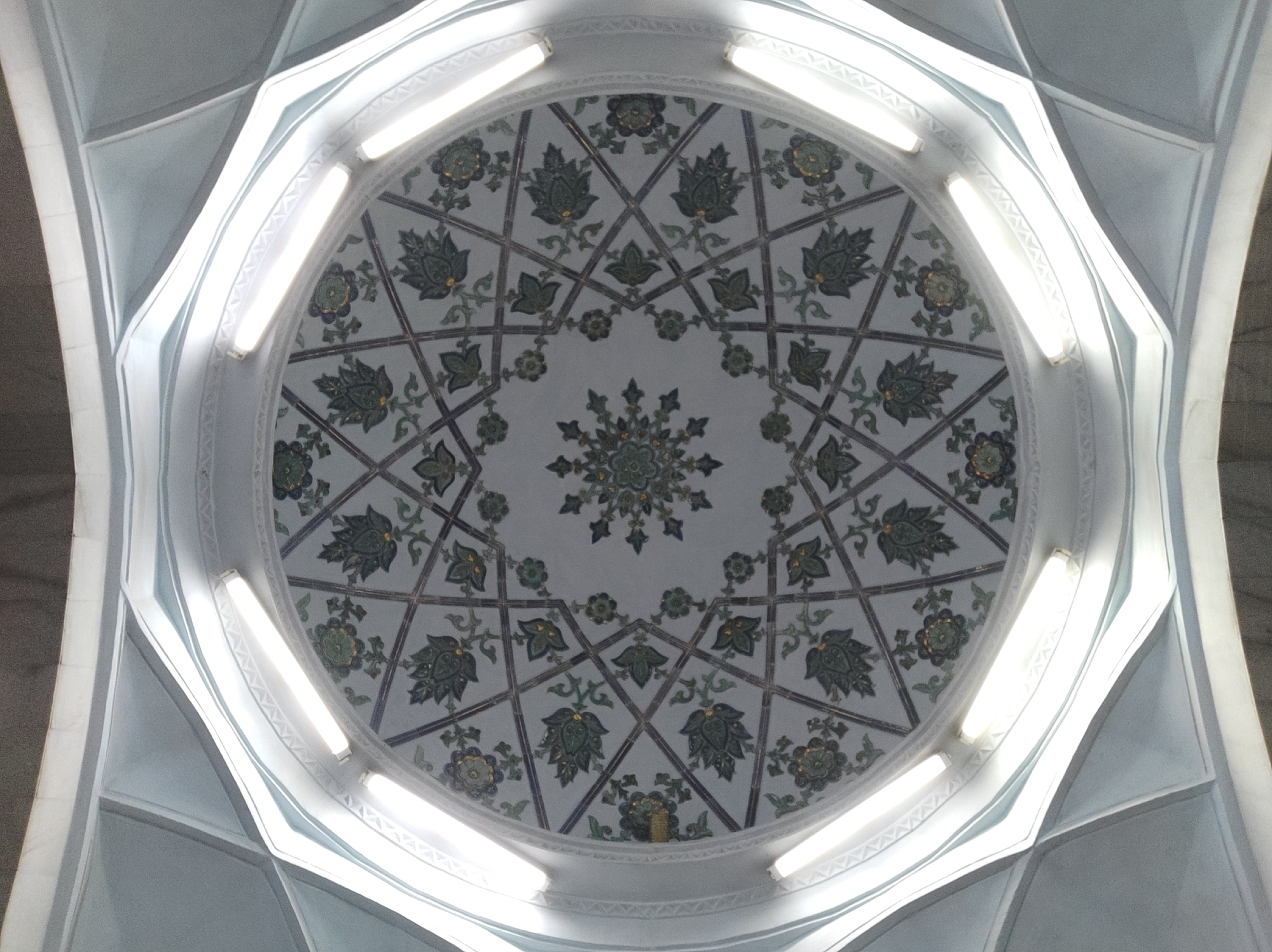 The image shows an interior view looking up at the ornate ceiling of a dome, likely a part of a religious or historical building. The dome's intricate design features a geometric star pattern with floral motifs. The patterns are predominantly in shades of green and blue on a white background. Linear fluorescent lights are embedded around the perimeter of the dome's interior, providing illumination. The architectural style suggests a Central Asian aesthetic, possibly from Uzbekistan. Given the architectural style and the mention of Tashkent, this is likely the interior of the Mirzo Ulugbek Madrasah in Tashkent, Uzbekistan, known for its distinctive dome and detailed tilework. No people are visible in the image.