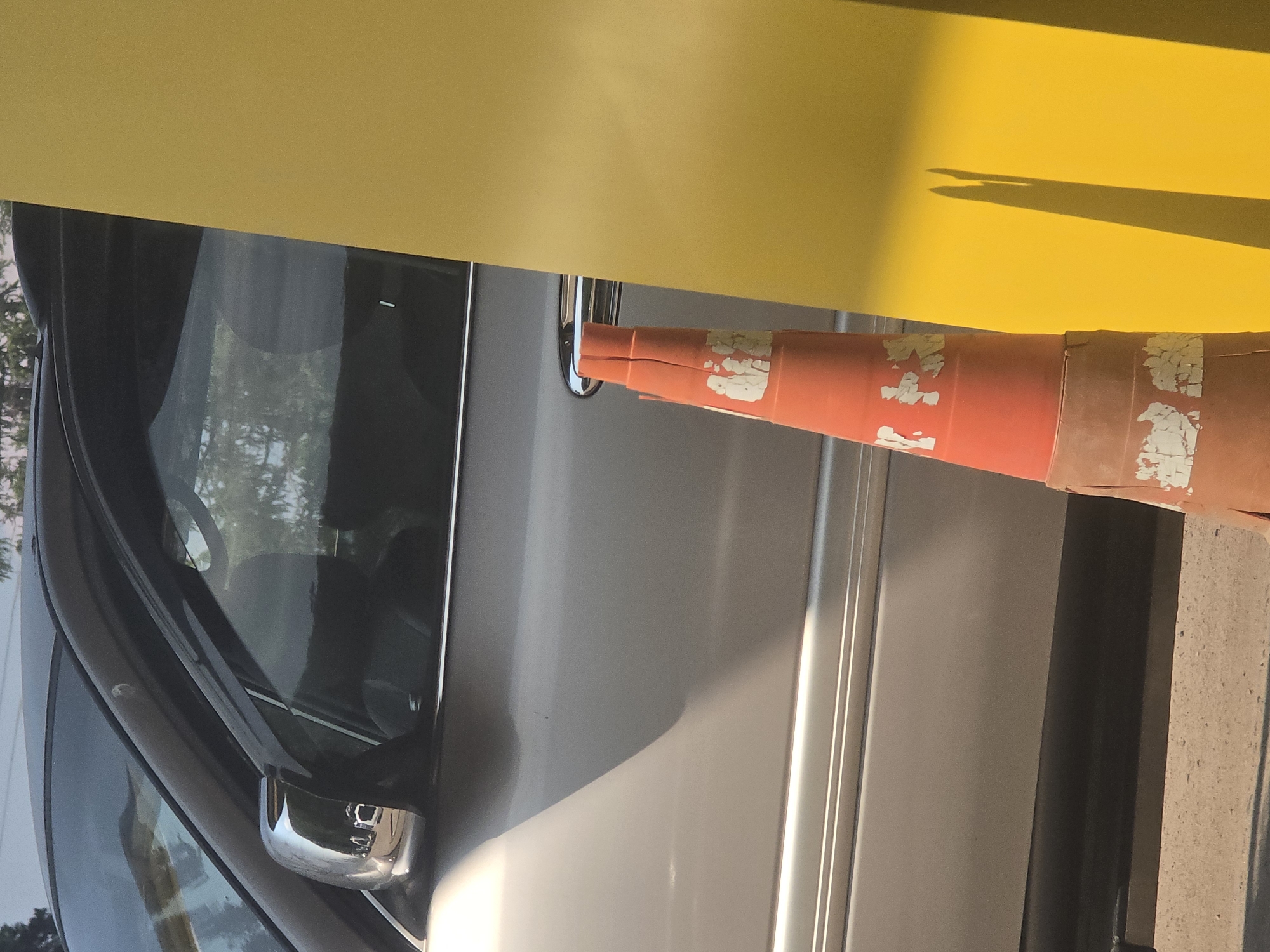 A traffic cone is positioned in front of a silver vehicle and a bright yellow vertical surface. The cone is orange with white peeling reflective tape. The vehicle's chrome side mirror is visible, reflecting other vehicles and trees. The scene appears to be an outdoor urban environment.