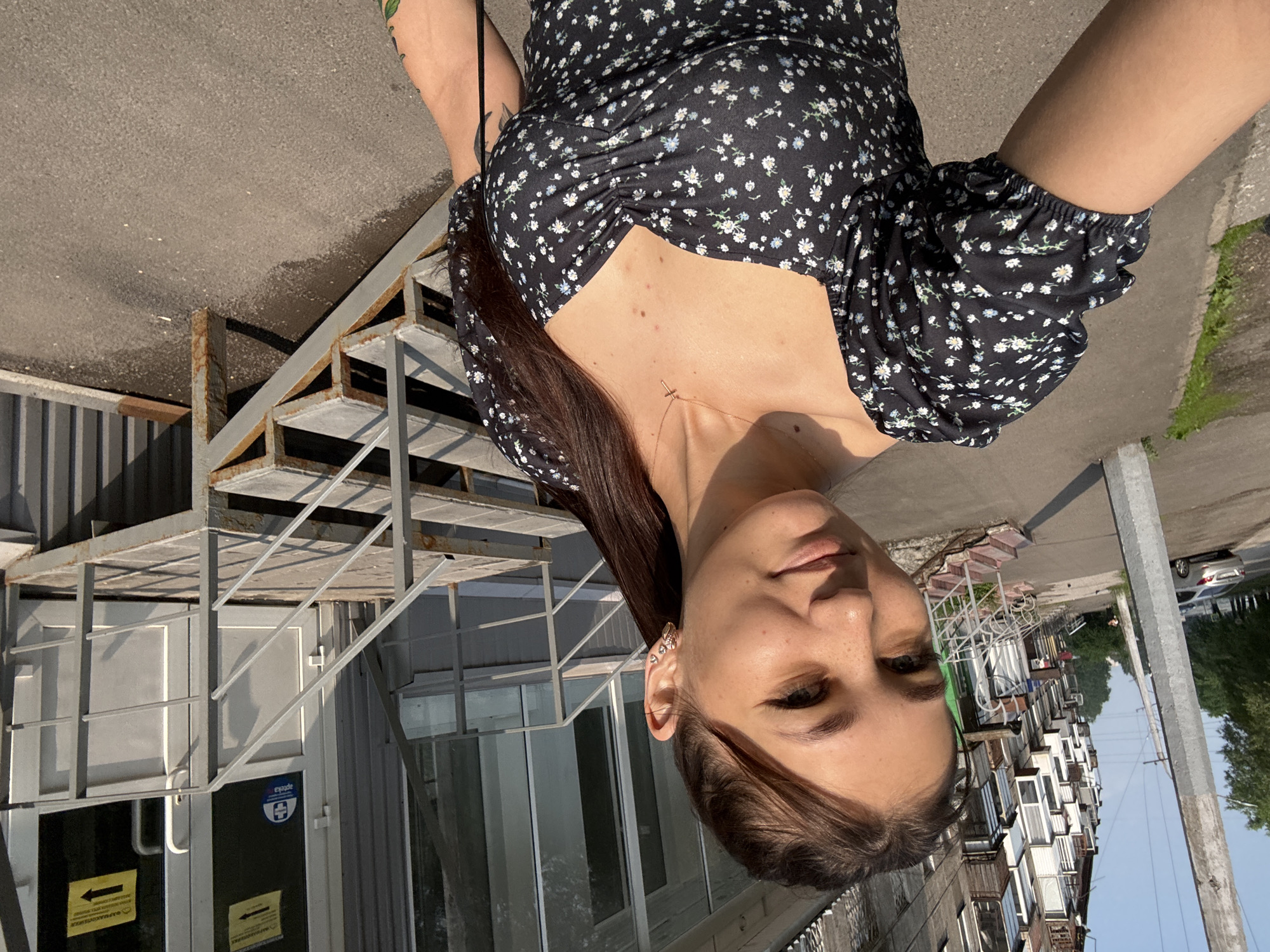 In Novokuznetsk, Russia, a young woman with brown hair pulled into a ponytail and a floral-print dress takes a selfie outdoors. She is standing on a sidewalk in front of a building with a metal staircase leading to an entrance. The building appears to be a commercial establishment, possibly a pharmacy, indicated by a blue circular sign with a white cross and the word "apteka" on the door. Yellow signs with black arrows are posted on the glass doors. The woman is looking at the camera and smiling. In the background, an apartment building with many balconies is visible. A car is parked on the street to the left of the frame. The lighting suggests it is a sunny day.