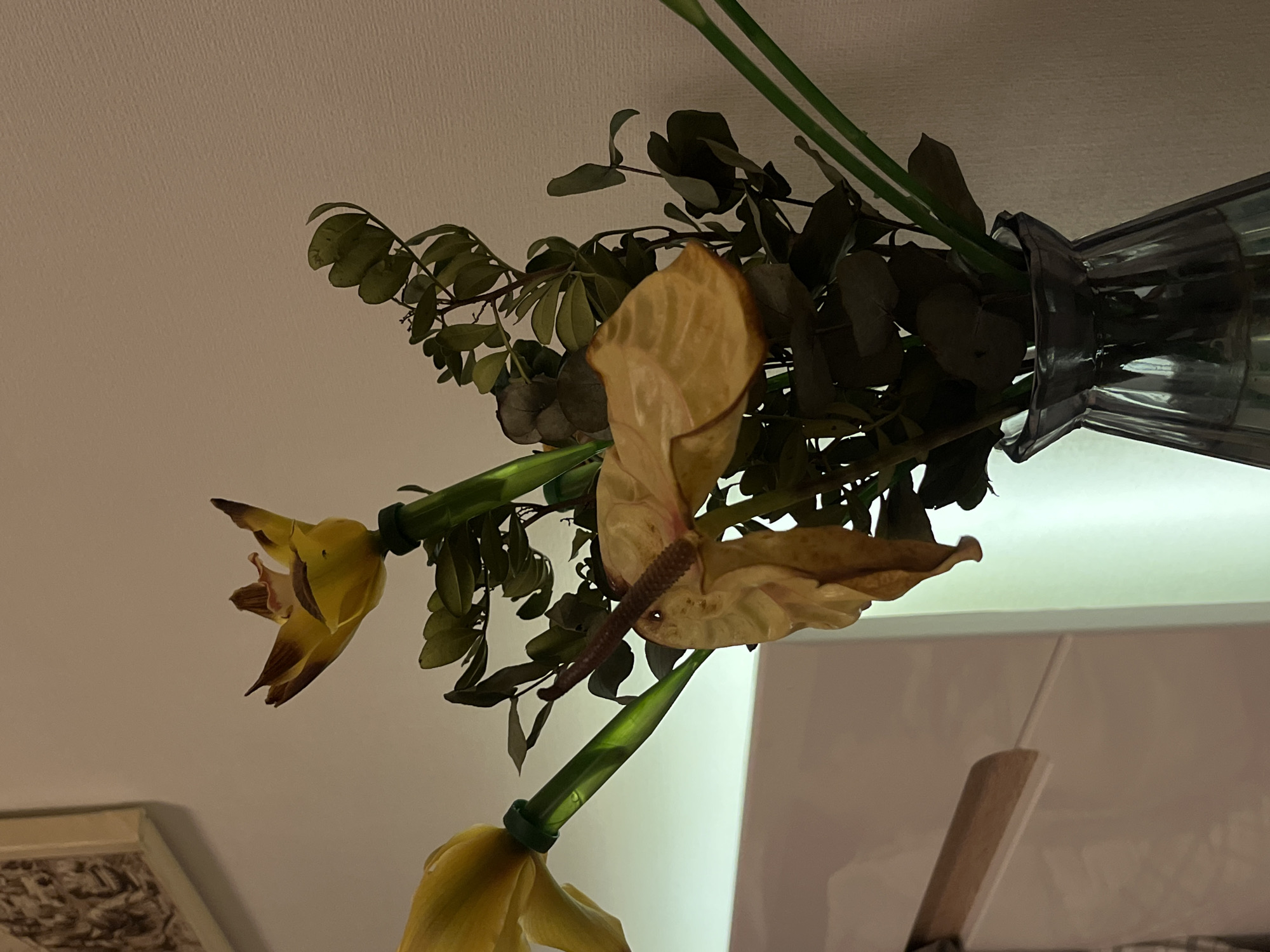 In an indoor setting, possibly a bedroom, a vase containing wilting yellow flowers and green foliage is placed on a surface. The flowers appear to be anthuriums and orchids, with some petals dry and curled. The green stems are visible, and some of the leaves are dark and dry, while others are still vibrant. The vase is made of dark, translucent glass with a ribbed texture and a flared opening. In the background, a textured white wall is visible, and to the left, a framed piece of art with a decorative pattern is mounted on the wall. A portion of a white piece of furniture with a wooden accent is also present in the lower left of the frame. The lighting in the scene is dim and warm, suggesting it might be evening or nighttime. There are no people or animals in the image.