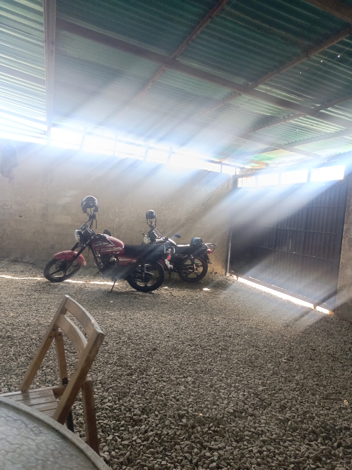 The scene is an indoor space in Juan Griego, Venezuela, with a roof made of corrugated metal allowing sunlight to penetrate through gaps. Two motorcycles are parked inside, each with a helmet resting on its handlebars. The ground is covered with gravel. A wooden chair is visible in the foreground. The background includes a wall and a dark metal gate, with sunlight streaming through the gate's openings. The image appears to be taken during a time of day when the sun is high, casting strong beams of light. There are no people visible in the image.