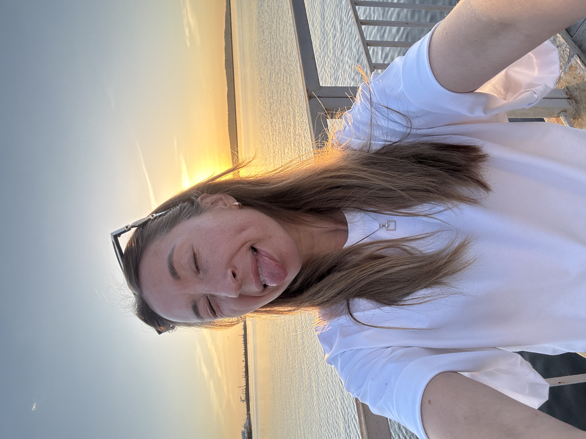 A woman is taking a selfie while standing on what appears to be a waterfront structure, likely a pier or bridge. She is wearing a white shirt and has sunglasses perched on her head. Her hair is light brown, and she is sticking her tongue out while winking one eye shut. The backdrop is a body of water reflecting a vibrant sunset. In the distance, there is what seems to be a cityscape with structures along the shoreline. Given the context of the city mentioned, this is likely the Kama River in Chaikovsky, Russia. A metal railing is visible to her right.