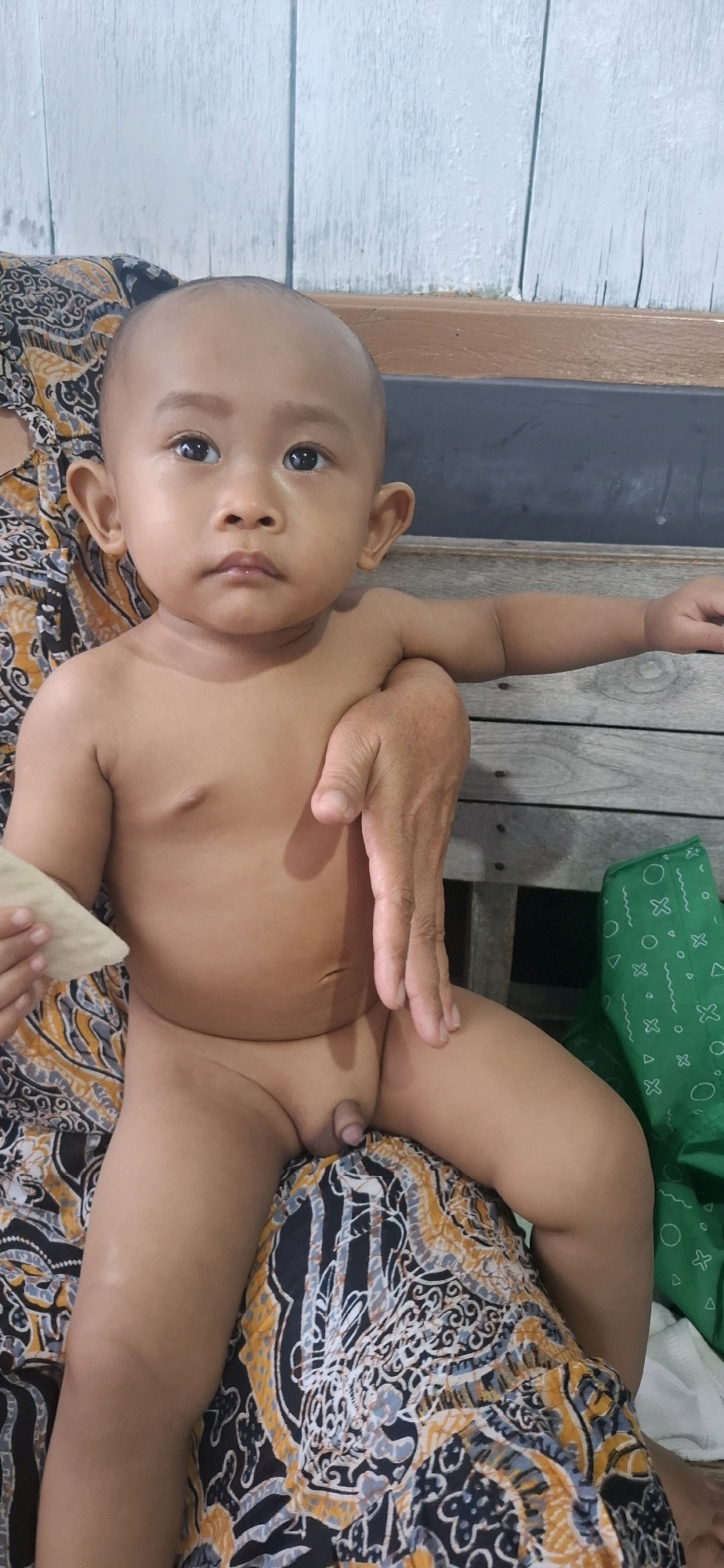 A child is sitting on a patterned fabric, possibly a sarong, with a person's hand resting on their chest. The child is unclothed and looking directly at the camera with a neutral expression. The background consists of a wooden wall with light blue paint and a wooden bench. A green fabric with white geometric patterns is visible to the right of the child. The setting appears to be indoors, in a casual environment, possibly a home in Desa Gagaksipat, Indonesia. The lighting suggests it is daytime. There is no visible text in the image.