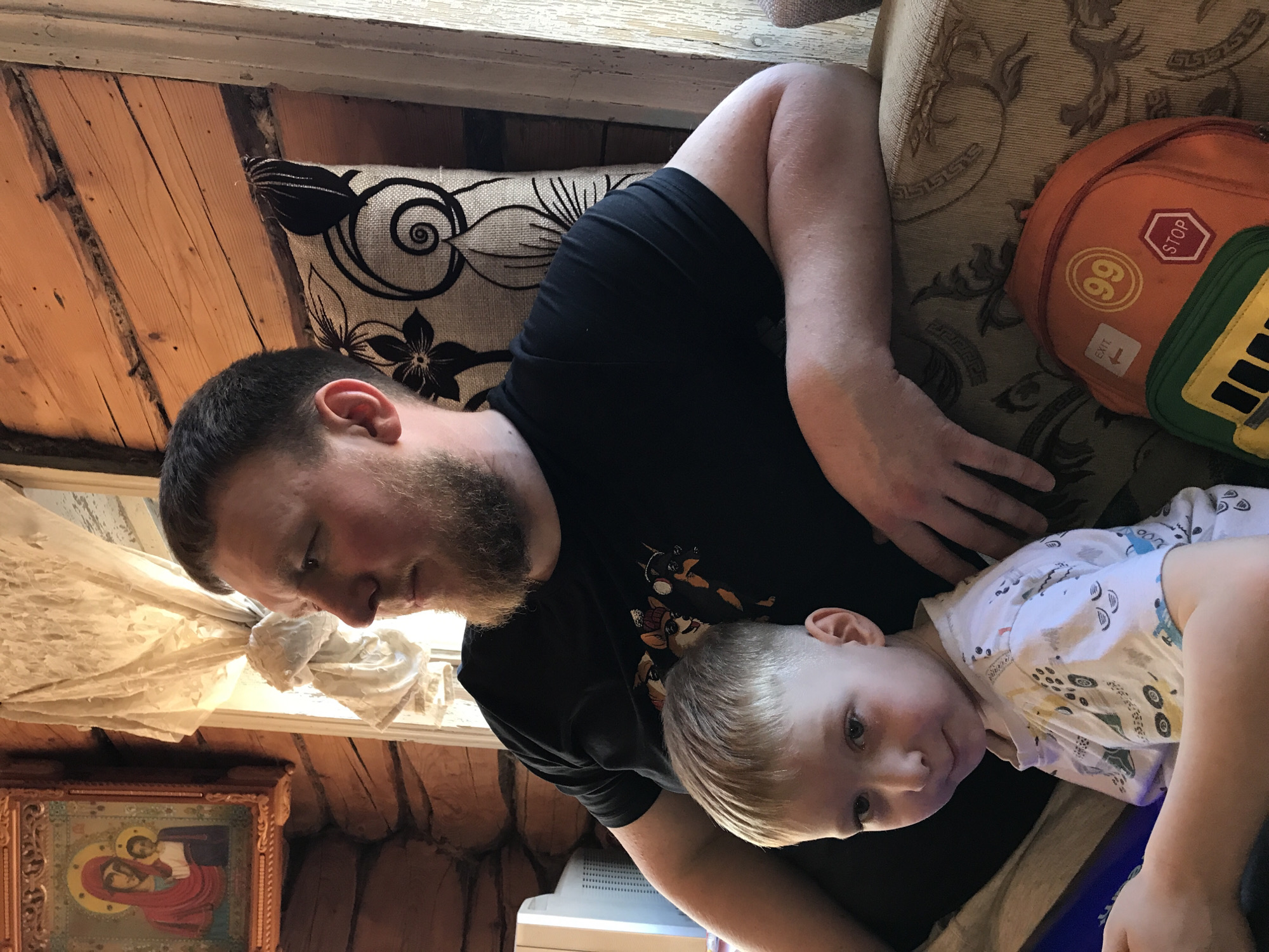 A man and a young child are indoors. The man has short brown hair and a beard, and is wearing a black t-shirt. He is lying down on a patterned sofa with a pillow. The child, with blond hair and wearing a white shirt with a pattern, is lying partially on the man. An orange and green backpack is on the sofa nearby. A window and a framed religious icon are visible in the background. The walls are constructed of logs. The location is Nizhnyaya Chermoda, Russia.