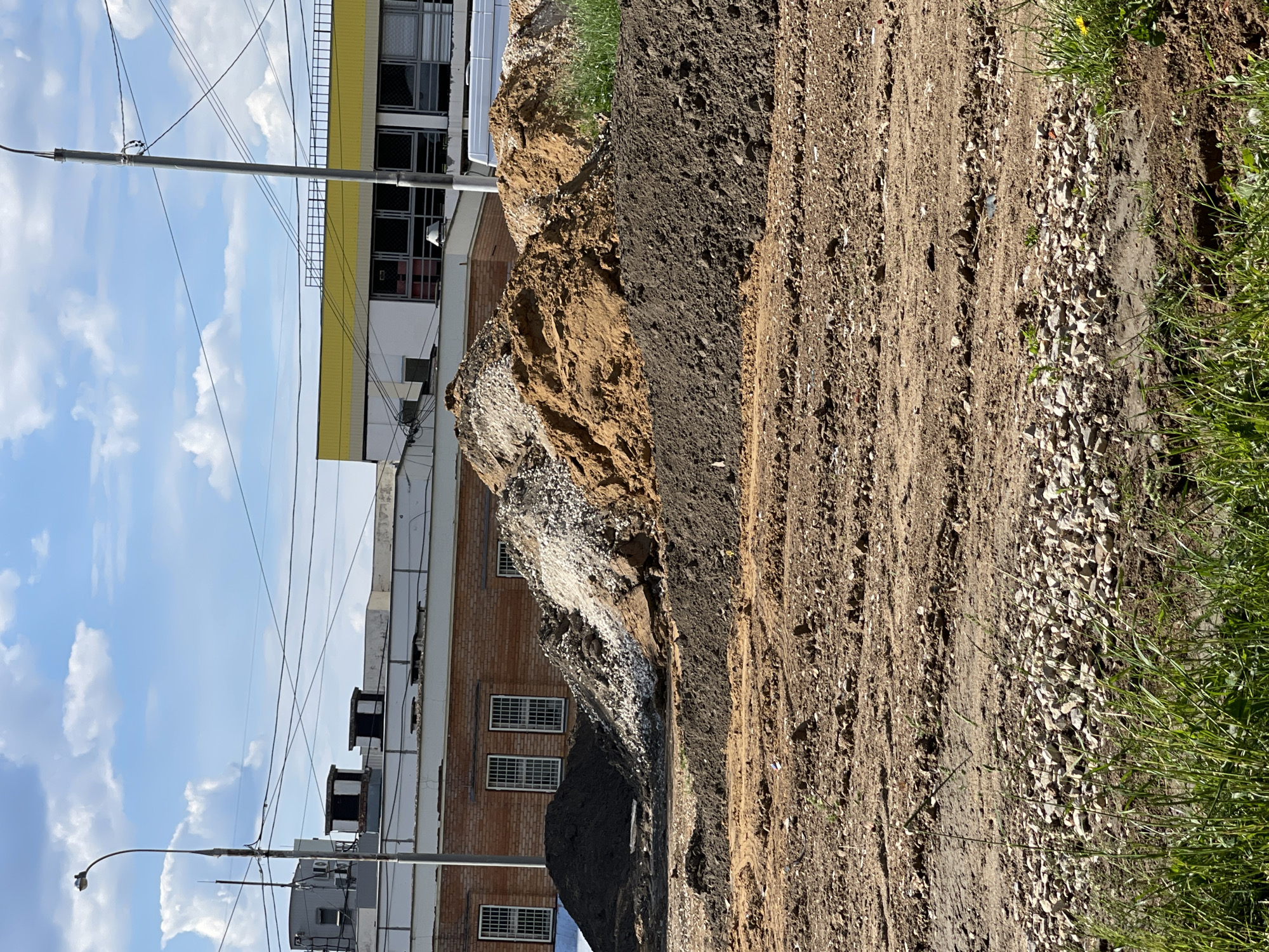 The image captures an outdoor urban scene in Moscow, Russia, depicting an area with significant ground disturbance, possibly related to construction or landscaping. The perspective is from ground level, looking towards some buildings under a clear sky.
In the foreground and midground, large piles of excavated material dominate the scene. To the left, a substantial mound of light brown earth is visible, partially covered with white and grey gravel or crushed stones. Adjacent to it, a separate pile of dark grey or black soil stands out. Further to the right, a wide, unpaved dirt path or road extends across the frame, showing tire tracks and interspersed patches of small white stones. A strip of vibrant green grass forms the rightmost edge of the frame, indicating the boundary of the disturbed ground.
In the background, on the left, two buildings are present. The more prominent one features a light-colored facade with a bright yellow trim along its upper level and numerous square windows with dark frames. Behind and slightly to its right, another building of reddish-brown brick construction with multiple windows is partially visible, obscured by the soil mounds. Above the buildings, utility poles with a complex network of power lines stretch across the frame, set against a bright blue sky with scattered white clouds. Various roof structures, including vents and air conditioning units, are discernible on the buildings.
The bright lighting and clear sky indicate that the photograph was taken during daytime with fair weather. No people or visible text are present in the image. The overall scene suggests an ongoing or recently completed earthwork project in an urban setting.