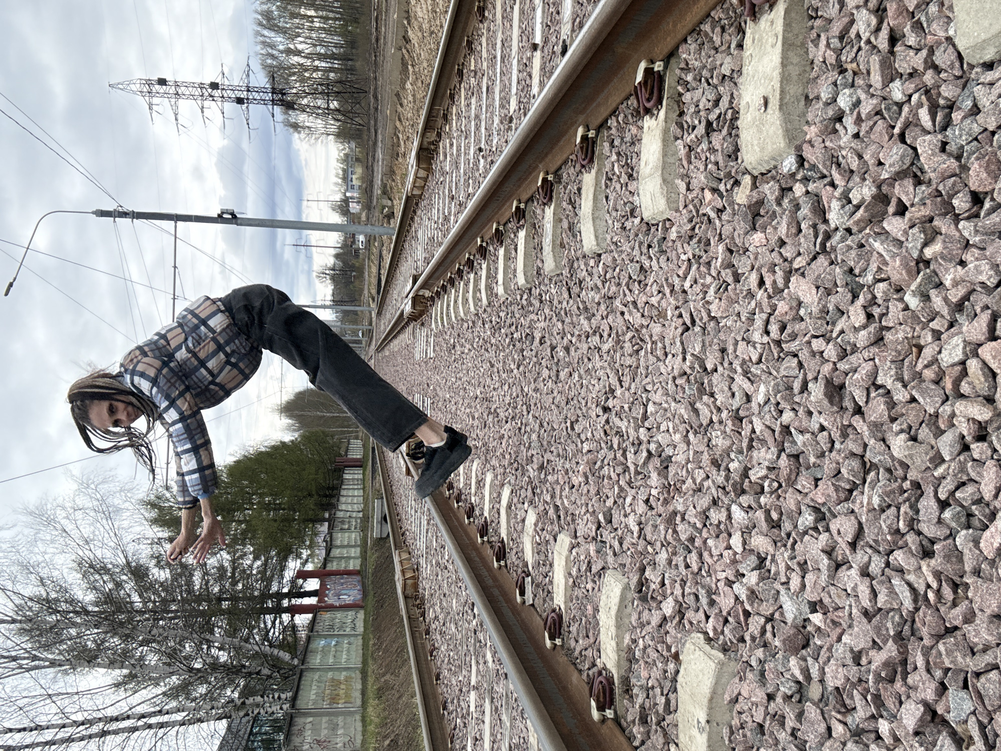 An outdoor image, rotated 90 degrees clockwise, captures a person striking a dynamic pose on a railway track in Rusanovo, Russia, under an overcast sky.
The main subject, a person with dreadlocks, is positioned with their body bent forward at the waist, arms outstretched, and one foot balancing on a rail. They are wearing a plaid shirt featuring brown, black, white, and light blue squares, dark pants, and dark shoes. Their posture suggests a moment of balance or a performance captured photographically.
The setting is a railway line featuring two sets of parallel tracks. The tracks are constructed with metal rails, concrete sleepers (ties), and a bed of pinkish-grey ballast stones. The tracks recede into the distance, converging towards a distant horizon.
In the mid-ground to the left of the tracks, a low, segmented fence or wall runs alongside, with some sections displaying what appears to be graffiti or artwork. Further back, a line of trees, some bare and some with nascent green foliage, borders the railway. A large metal lattice electricity pylon stands prominently against the cloudy sky in the background, accompanied by a utility pole with overhead wires and a street light fixture closer to the foreground. Distant, indistinct buildings are visible on the horizon.
The weather is cloudy or overcast, providing soft, diffused light, indicating daytime. There is no discernible text visible in the image.