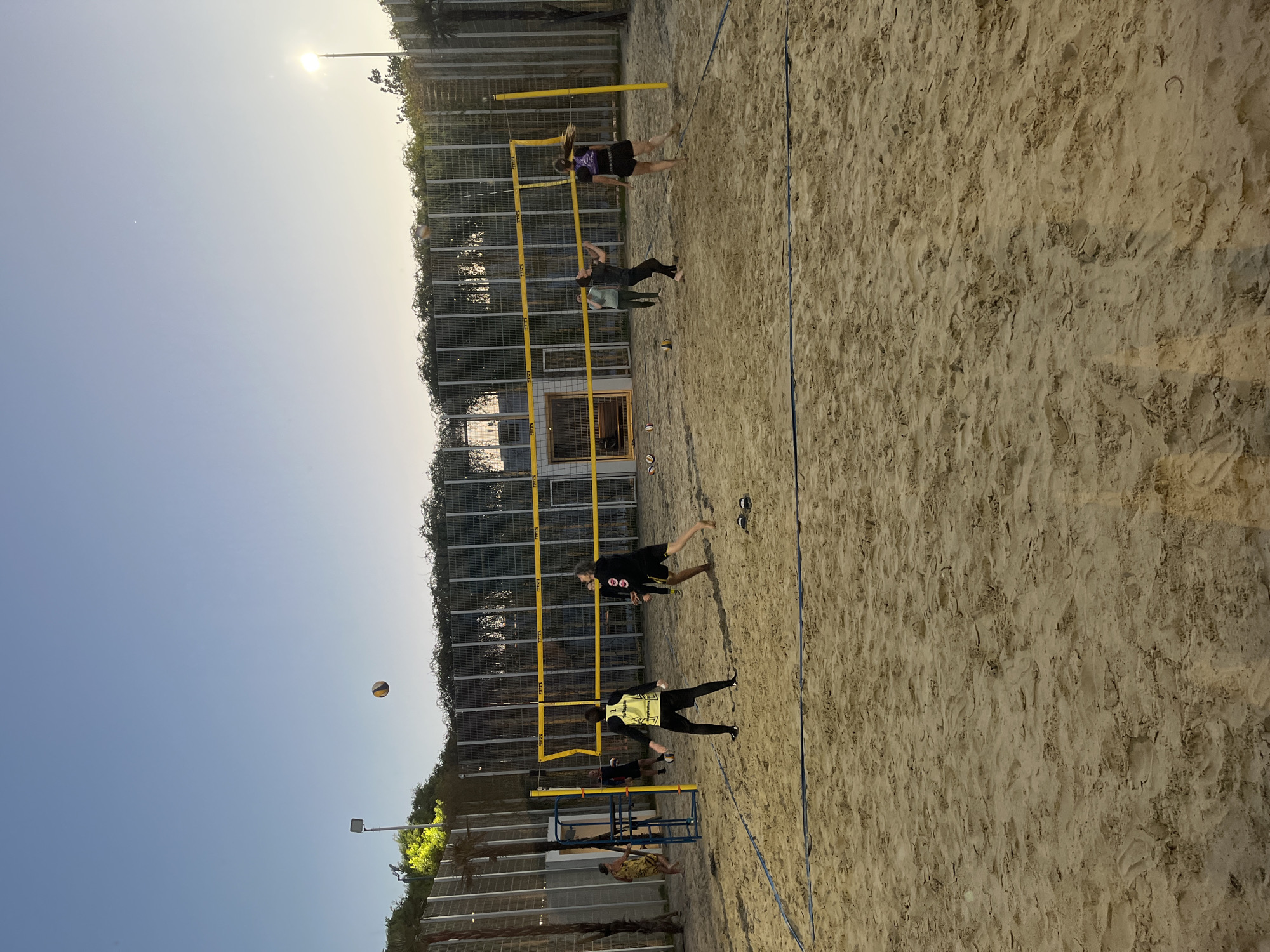 The image depicts a beach volleyball game in progress on a sand court. There are at least six people visible, actively involved in the game. One individual is airborne, presumably spiking or hitting the ball over the net. Others are positioned on the court, seemingly ready to receive the ball. The volleyball is visible in the air. The net is standard yellow and blue color of the mesh. The court is surrounded by a fence with foliage growing on it. The location is likely in Krasnodar, Russia based on the image's geotag.