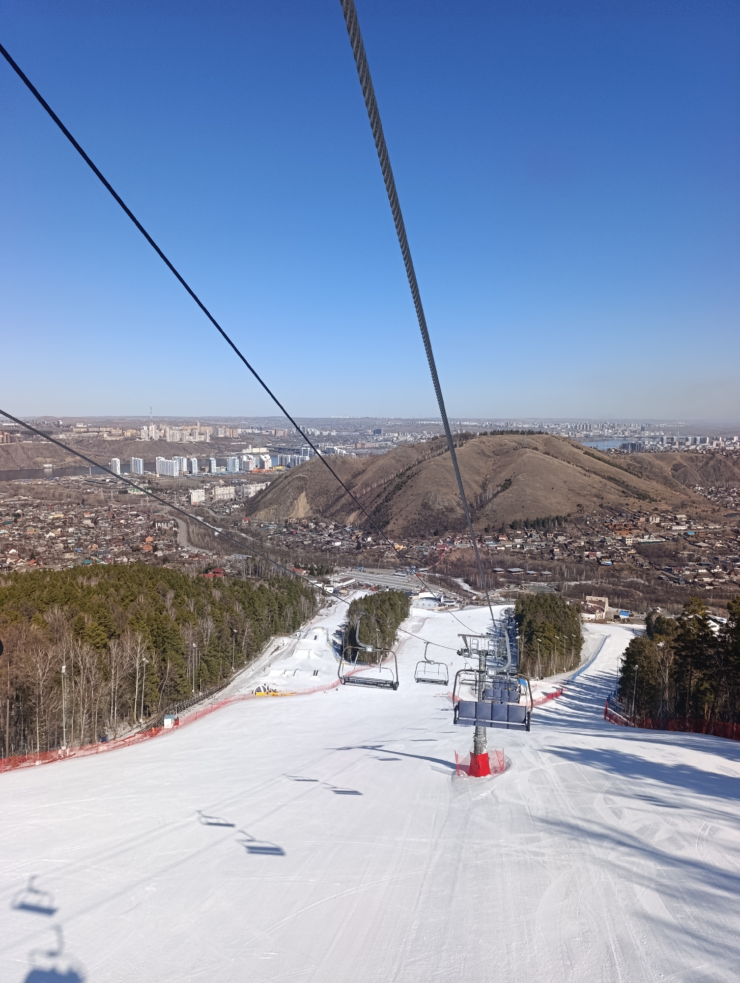 The image depicts a ski slope at the Bobrovy Log Fun Park in Krasnoyarsk, Russia. The view is from a ski lift, looking downhill. The slope is covered in snow and groomed with visible tracks. Several empty ski lift chairs are visible along the cable. A safety fence is visible on both sides of the slope. Beyond the slope, the city of Krasnoyarsk stretches out with buildings and the Yenisey River visible in the distance. A large, mostly bare hill rises behind the urban area. The sky is clear and blue.