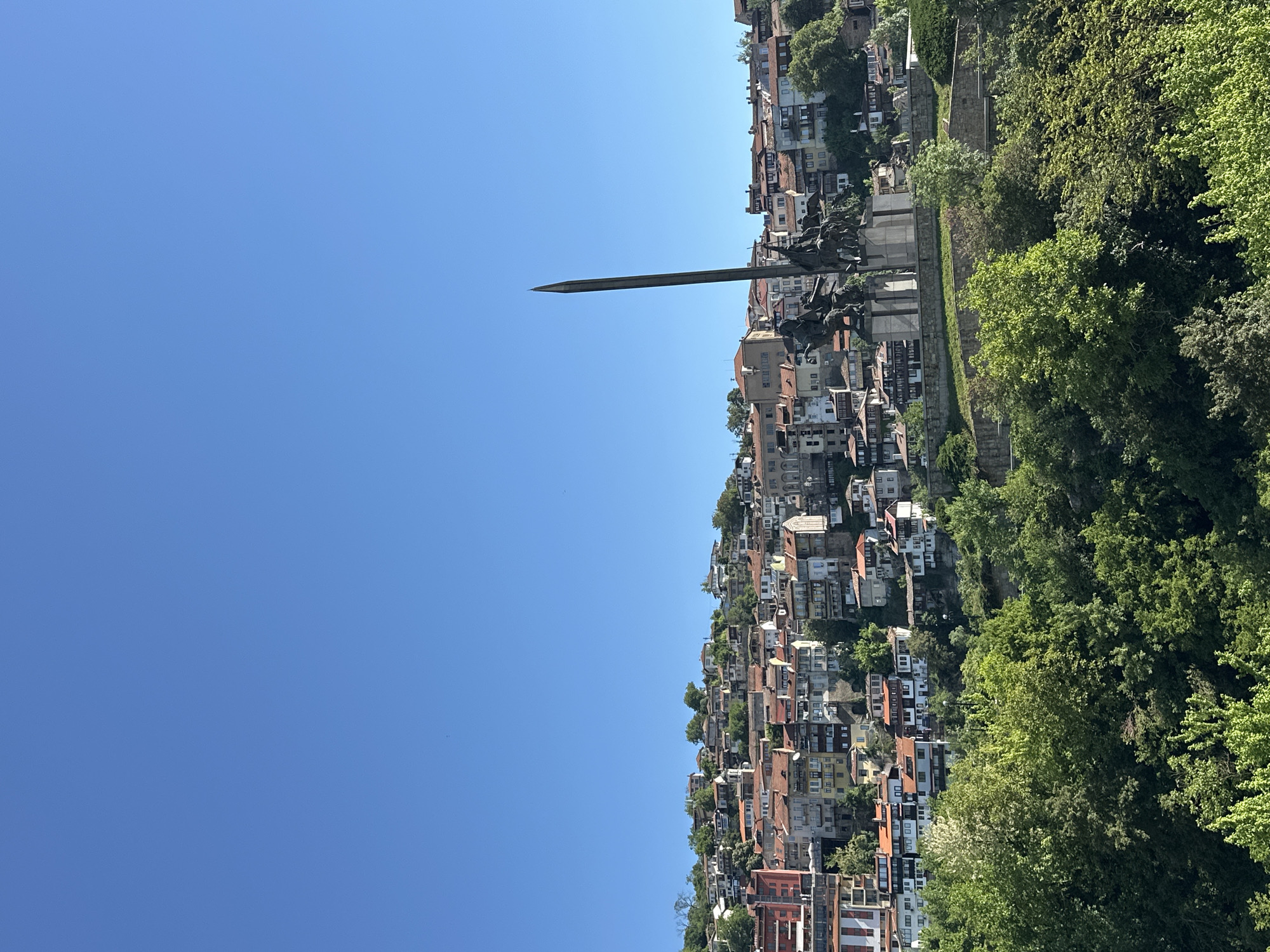 The image shows a view of Veliko Tarnovo, Bulgaria, featuring a dense cluster of buildings and a prominent monument with a tall, thin, sword-like element pointing into the sky. The buildings vary in color and style, covering the hillside. To the right, lush green trees grow along the hillside. A clear blue sky fills the majority of the frame. The angle suggests the photo was taken facing up towards the monument.