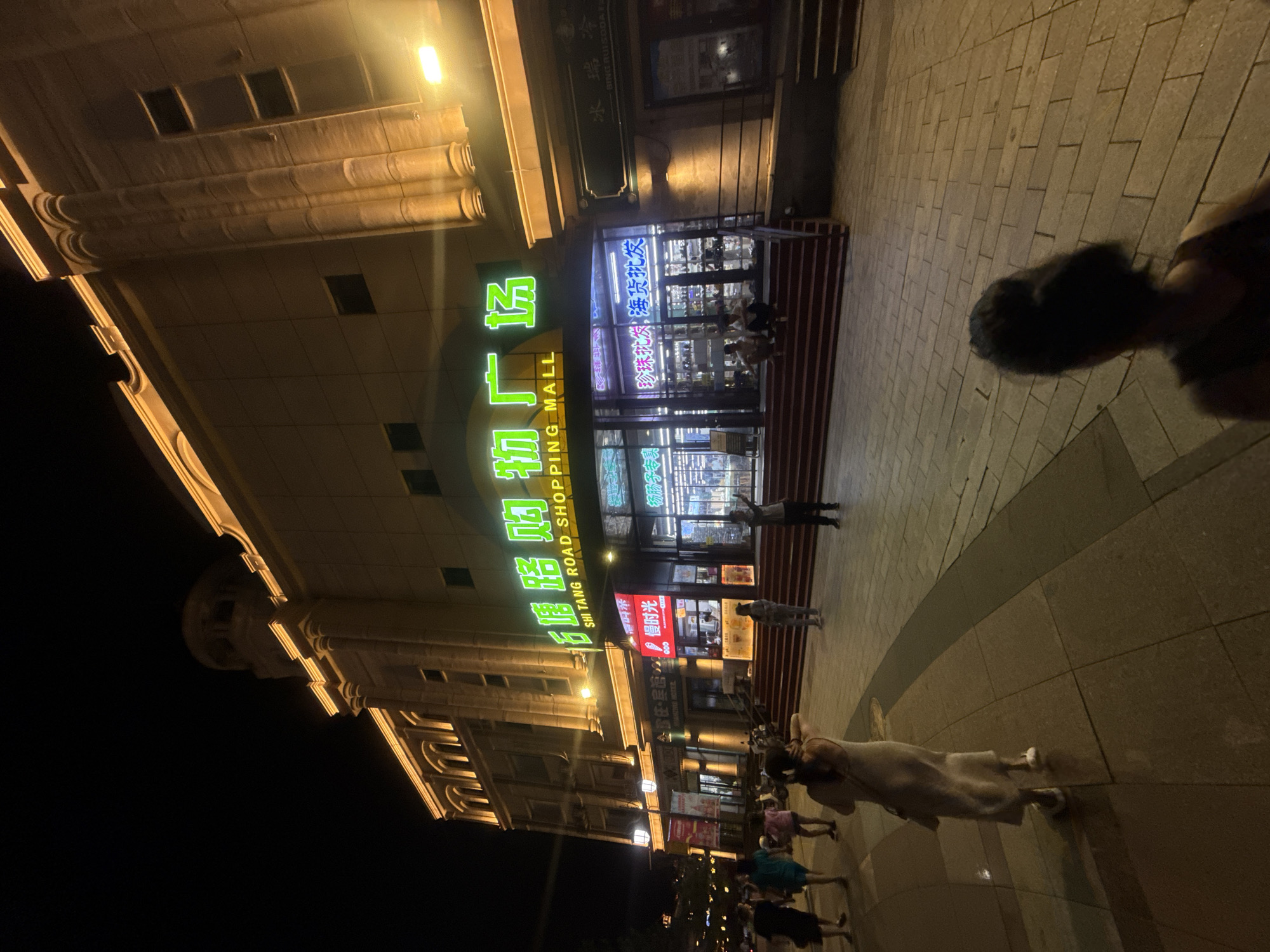 This is a nighttime, wide-angle, eye-level photograph taken on a sidewalk in front of the Shi Tang Road Shopping Mall in Qinhuangdao, China. The mall's facade is illuminated with warm yellow lights along its upper cornices and columns. A prominent, bright green neon sign above the entrance reads "石塘路购物广场" (Shi Tang Road Shopping Mall). The mall's entrance features red steps leading up to glass doors, with several illuminated signs visible inside.
Several people are present on the sidewalk and steps. In the foreground, the back of a woman in a light-colored dress is visible, walking away from the camera. Further back, a group of people are walking on the sidewalk, and two individuals are seated on the red steps of the mall entrance. Another person stands near the mall entrance, appearing to pose for the photo. The sidewalk is paved with light-colored square tiles, and a darker curved section separates it from a paved area in front of the mall. The overall scene depicts a commercial area at night, with shoppers and passersby.