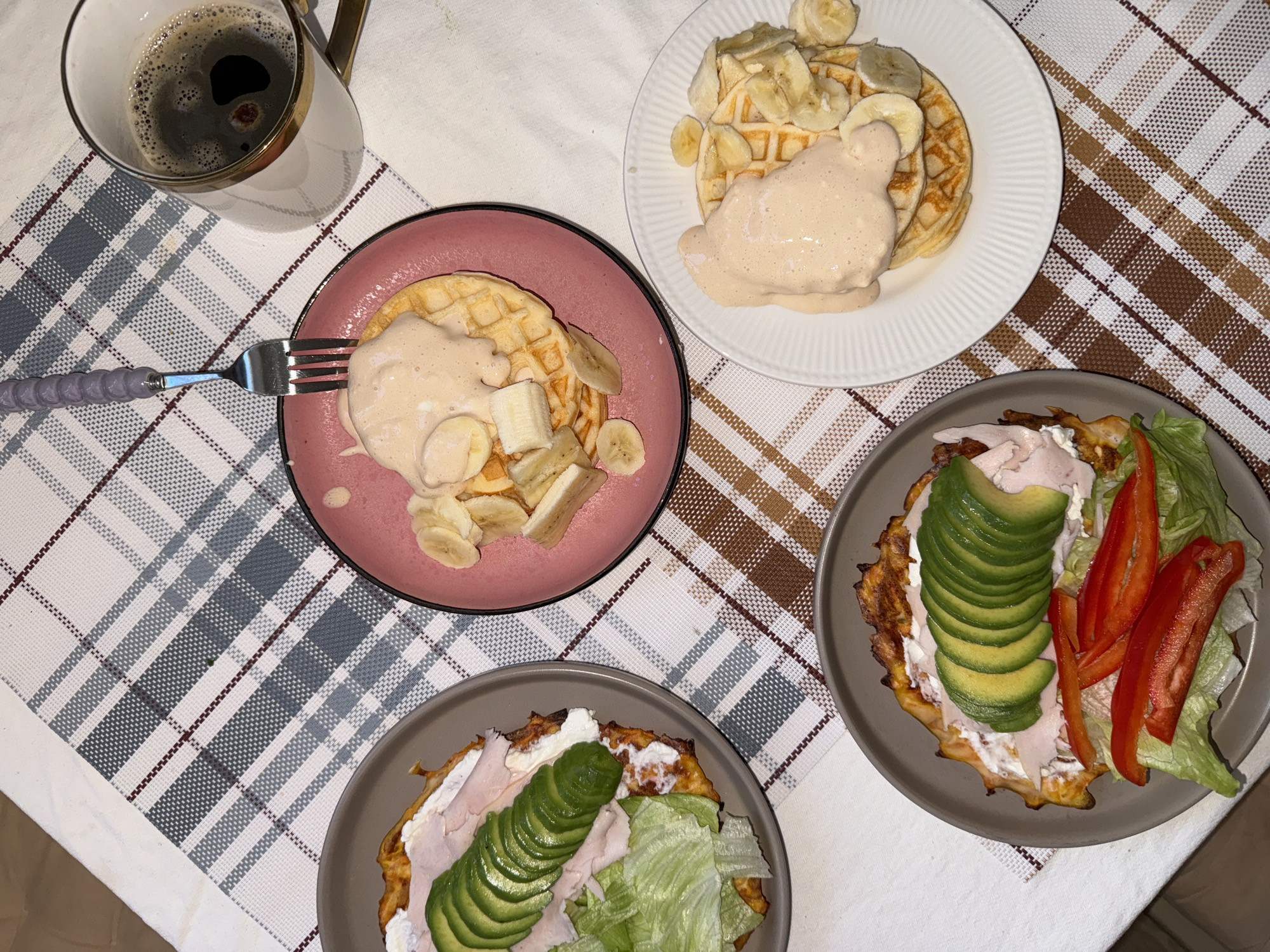 The image shows a breakfast scene with multiple plates of food and a mug of coffee arranged on a tablecloth. Two plates contain waffles topped with banana slices and a light-colored sauce. The other two plates feature a waffle base topped with cream cheese, sliced avocado, and, on one, red bell pepper slices and lettuce. One plate has turkey slices and lettuce on top. A fork with a purple handle is on the left of the waffle plate. The mug of coffee is near the top left of the image. The tablecloth has a checkered pattern. There is no indication of Krasnoyarsk, Russia, or a specific location in the image.