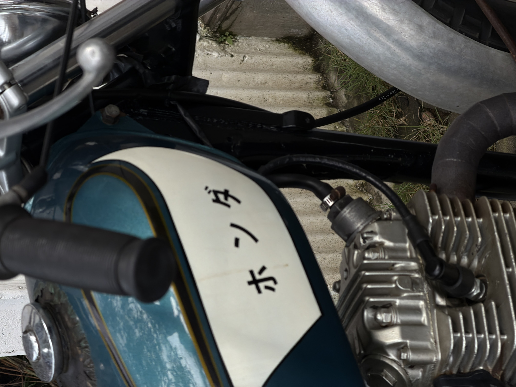 The image shows a close-up view of a vintage motorcycle, likely in a residential setting given the weathered concrete wall and overgrown grass in the background. The motorcycle's teal-colored fuel tank has a white decal with Japanese characters that read "ホンダ" (Honda). A chrome gas cap is visible on the left side of the tank. Part of the engine, including fins and a spark plug wire, is in the foreground. A silver fender curves into the right side of the frame. The scene appears to be outdoors, possibly in a driveway or yard, in Mengwi, Indonesia. There are no people visible in the image.