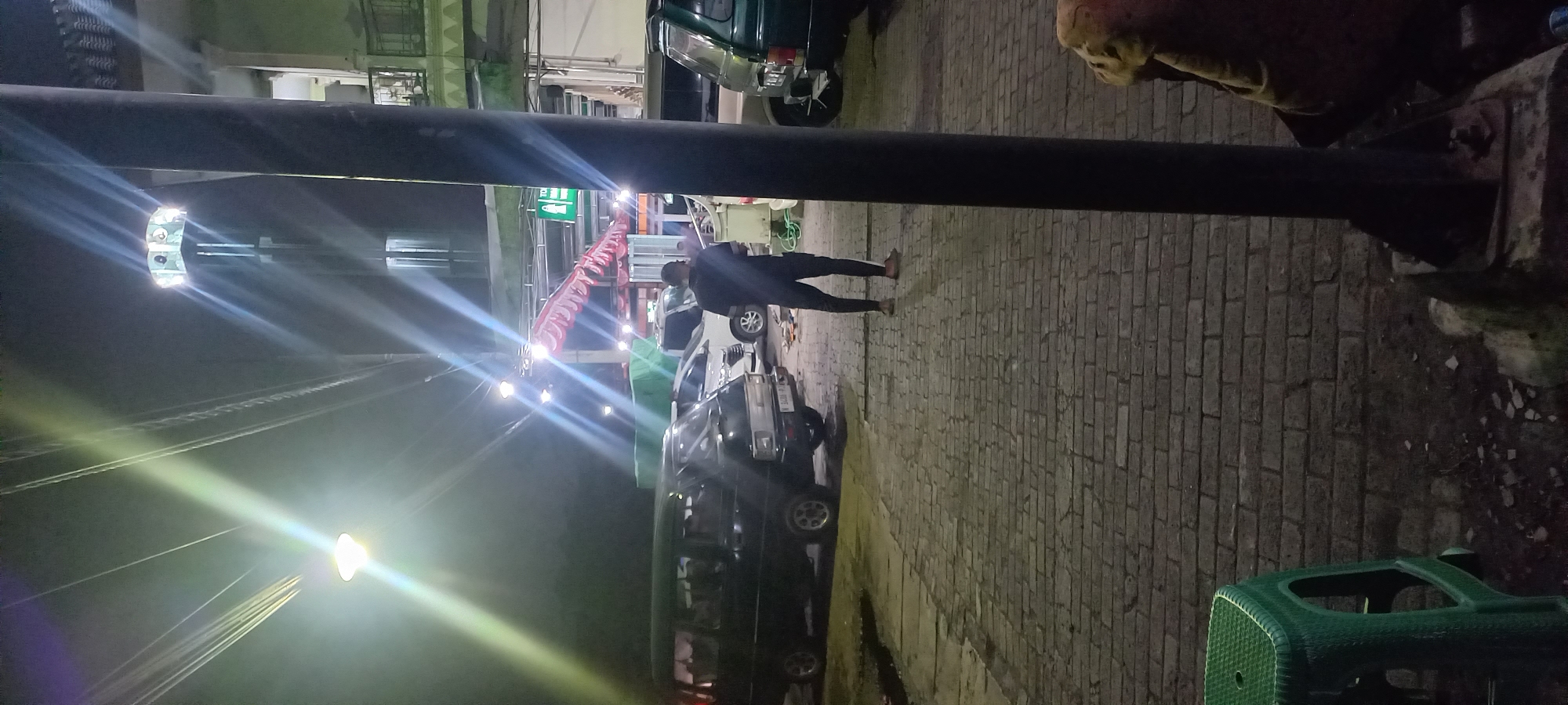 The image depicts a nighttime street scene in Banjarsari, Indonesia. The foreground is dominated by a cobblestone pavement and a green plastic stool. To the left, a black van is parked. Further back, several other vehicles are visible, including a white car and parts of other vans. A man wearing dark clothing and a cap is standing in the middle ground, facing away from the camera. He appears to be holding something in his hands. In the background, buildings with illuminated windows and balconies are present, along with streetlights and awnings. The overall lighting suggests artificial illumination from streetlamps and building lights, creating a slightly hazy atmosphere.