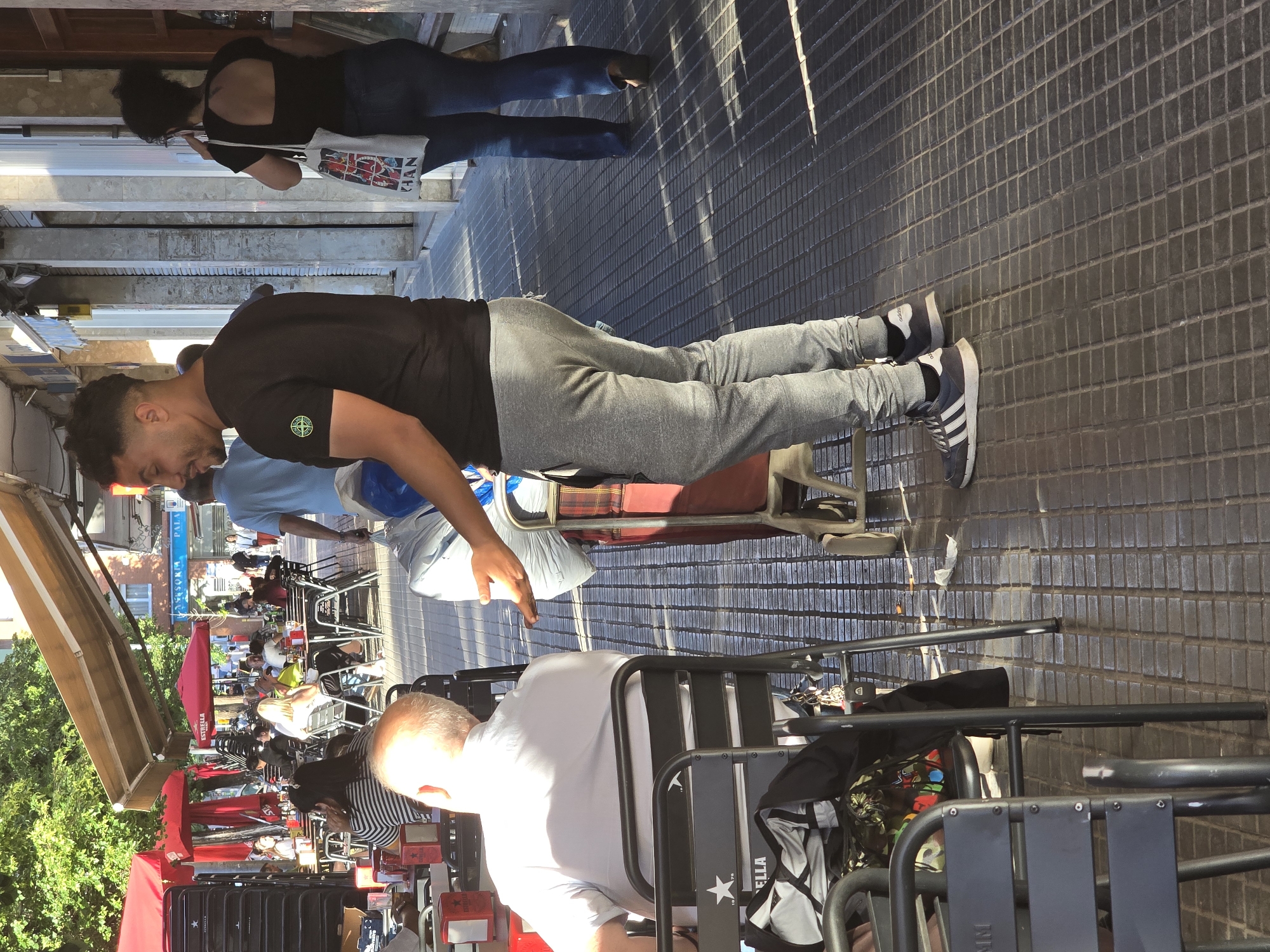 The image shows a street scene with a row of outdoor cafe tables and chairs, densely packed with patrons. The scene appears to be in Cornellà de Llobregat, Spain, as evidenced by the "Palasor" sign visible. The setting is outdoors on a sunny day, with shadows cast on the paved street.
In the foreground, a bald man in a white shirt is seated at a table. A stack of chairs with Estrella branding is visible near him. A man wearing a black t-shirt and grey sweatpants is standing behind the seated man, gesturing with his right hand. He is wearing Adidas sneakers.
Further down the row of tables, people are seated and interacting with each other, creating a bustling social atmosphere. Red awnings and trees provide shade for the patrons. In the background, a woman with dark hair is walking away from the scene on the sidewalk, wearing a black tank top and jeans. She has a white bag on her shoulder.