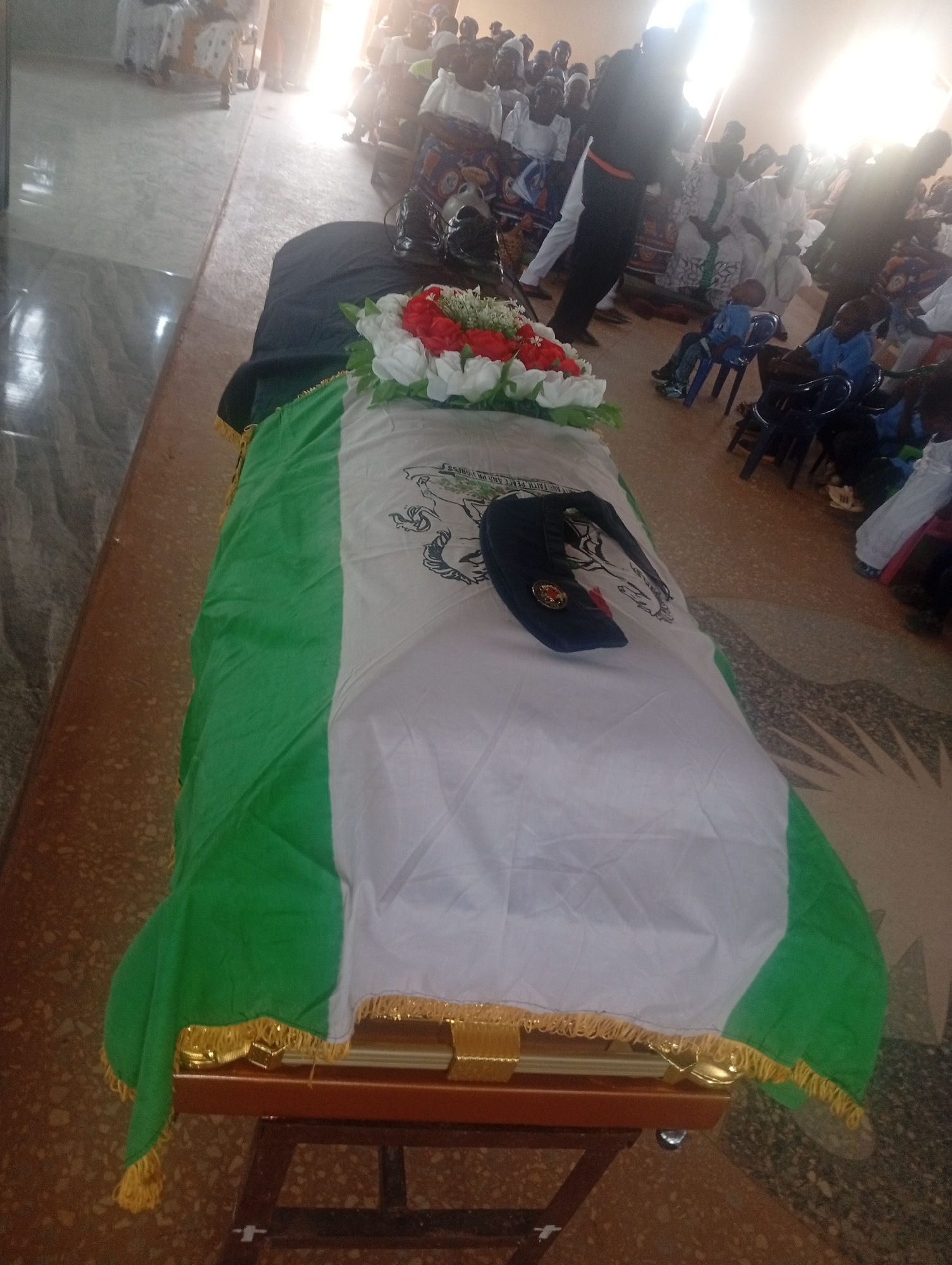 The image shows a funeral in a building with a patterned tile floor. A coffin draped with a green and white flag is prominently displayed in the foreground. A floral arrangement with red and white flowers sits on top of the coffin, along with a dark-colored hat. People are seated in the background, attending the service. The location is Unknown, Unknown.