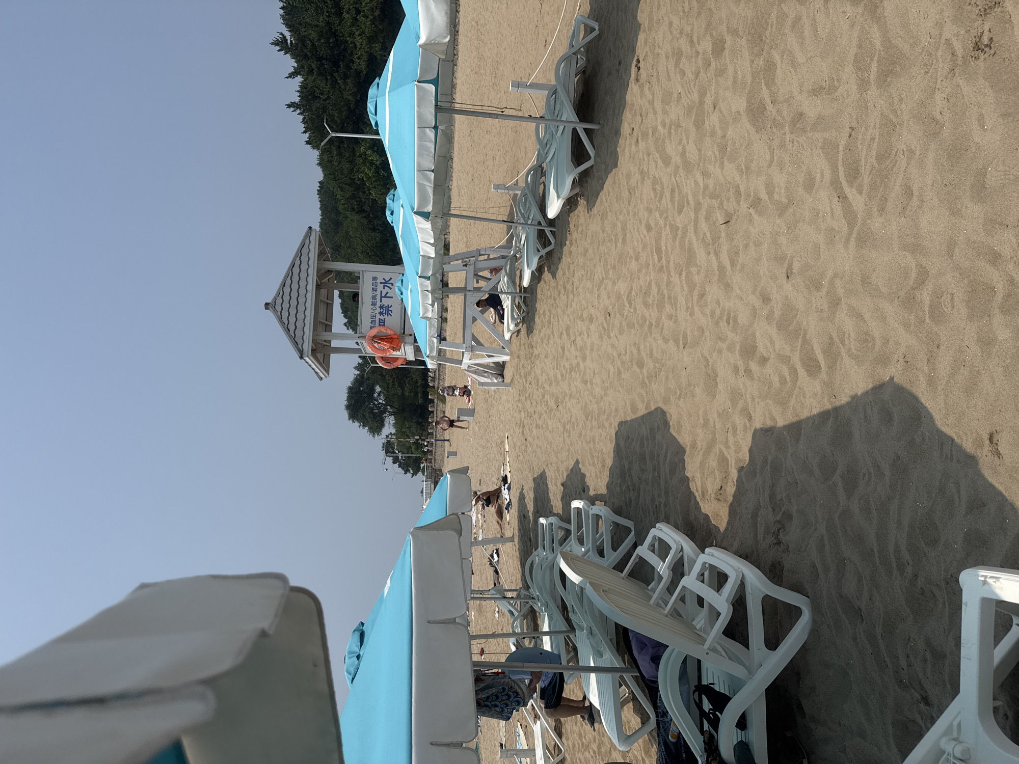 The image depicts a sandy beach on a sunny day, likely a section of Beidaihe Beach in Qinhuangdao, China, known for its tourist resorts. In the foreground, several white plastic lounge chairs are arranged under blue and white beach umbrellas, some with people partially visible. To the right, a lifeguard tower stands prominently, with an orange lifebuoy attached and signage in Chinese characters indicating restrictions on swimming for individuals with certain health conditions. Further back, other beachgoers are seen scattered across the sand, some sunbathing and others walking. The background features a line of green trees and a clear blue sky. The overall scene is tranquil and suggestive of a typical summer day at a coastal resort.
