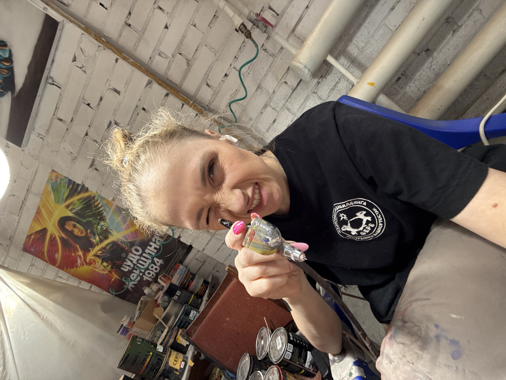 An indoor environment in Chaikovsky, Russia, is depicted, resembling an art studio or workshop. A blonde-haired female subject, with hair gathered in a bun, is positioned mid-shot, looking directly at the camera with a squinting, smiling expression. She wears a black short-sleeved t-shirt featuring a white circular logo on the left chest, which displays text including "ФЕДЕРАЦИЯ ПЕЙНТБОЛА", "Г. ЧАЙКОВСКИЙ", and "РЕСПУБЛИКИ БАШКОРТОСТАН". A white wireless earbud is visible in her right ear. Her right hand, adorned with pink nail polish, holds an airbrush coated with multi-colored paint, pointing towards the camera. She wears light-colored trousers, which show evidence of paint splatter. The background consists of a white brick wall with exposed utility piping, including a green flexible pipe and several off-white pipes. A "Wonder Woman 1984" movie poster, bearing the text "ЧУДО ЖЕНЩИНА 1984" in Cyrillic, is mounted on the wall. To the subject's left, shelves are stocked with numerous paint cans, one of which is labeled "CERTA". A maroon piece of furniture is partially visible behind her left shoulder, and a blue object below her right arm. Overhead lighting illuminates the scene, and a white sheet or tarp hangs from the ceiling on the left.