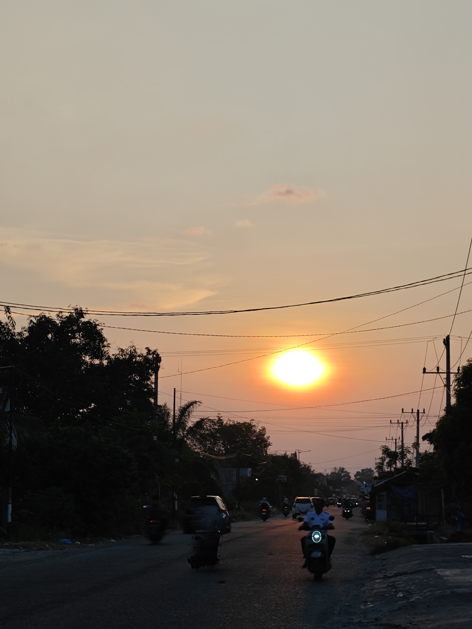 The image depicts a street scene in Blangcot Tunong, Indonesia, at dusk with the sun low on the horizon. A large, intensely bright, circular orange-yellow sun dominates the sky, positioned slightly left of center above the distant tree line. The sky exhibits hues of orange and yellow near the horizon, transitioning to lighter tones higher up, with minimal cloud cover.
A two-lane asphalt road extends from the foreground into the background, where traffic flows in both directions. In the foreground, a motorcycle with a prominent circular LED headlight is ridden by an individual wearing a light-colored, possibly white, collared shirt, facing away from the viewer. Behind this motorcycle, several other vehicles, primarily motorcycles with illuminated headlights and taillights, are visible traveling in both directions, appearing as silhouettes against the setting sun or as bright points of light. A light-colored car is also discernable further down the road on the left, and a dark-colored van-like vehicle is moving away on the right.
The left side of the road is bordered by a dense, dark mass of trees and foliage. Overhead utility poles with multiple crisscrossing wires are visible above the tree line on both sides of the road. On the right side, the environment appears less densely vegetated, with a small, dark structure, possibly a shack or vendor stall, topped with a blue tarpaulin roof, situated near the road. The immediate ground on the right shoulder appears unpaved and somewhat uneven. The overall scene is characterized by the low, warm light of the setting sun, casting significant portions of the foreground and roadside elements into silhouette.