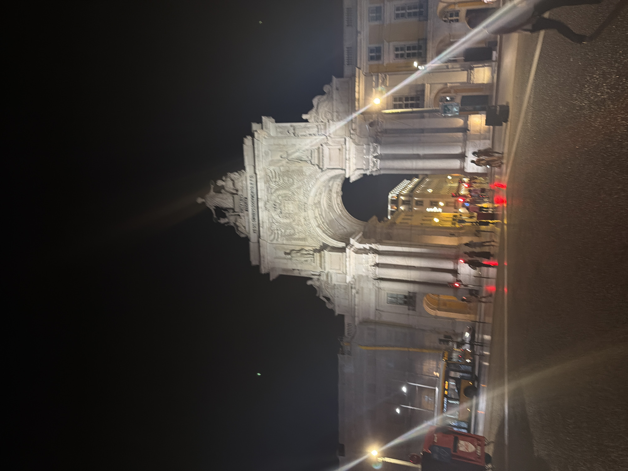 This is a night-time, eye-level, slightly low-angle, long-shot, vertical photograph taken in Praça do Comércio, Lisbon, Portugal. The main subject is the Arco da Rua Augusta, a triumphal arch adorned with sculptures and inscriptions, illuminated by artificial lights. The archway frames a view of buildings in the distance. In the foreground, a street scene unfolds with a yellow bus on the left side of the frame and several pedestrians walking on the wet street. Red traffic lights are visible. The sky is dark, with a few faint streaks of light from artificial sources. The overall atmosphere is that of a bustling city square at night.
