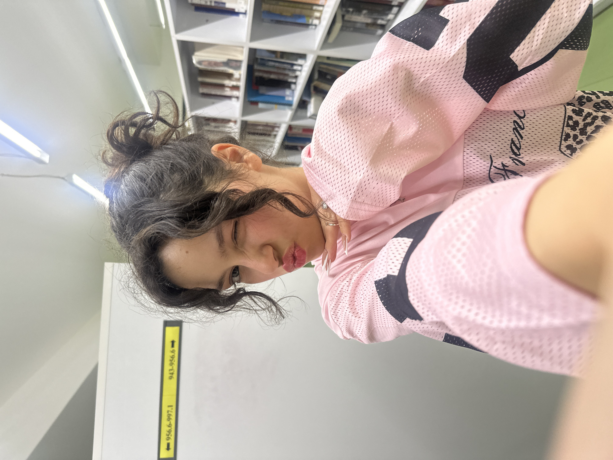 The image displays a selfie of a young female taken inside a library in Yerevan, Armenia. The subject, centrally framed, has dark, curly hair styled in a messy bun. She is winking with her left eye and puckering her lips. Her right hand, featuring a silver-colored ring on the ring finger and long, pointed light-colored fingernails, rests beneath her chin. She wears a pink, mesh-textured long-sleeved top with black geometric patterns on the sleeves and a leopard print section with partially visible cursive text "Franc" on the torso. The background on the left consists of a light-colored wall with a yellow directional sign displaying "← 956.6-997.1" and "943-956.6 →", indicating Dewey Decimal Classification ranges. To the right, white bookshelves are filled with numerous books. Overhead, linear fluorescent lights illuminate the indoor setting.