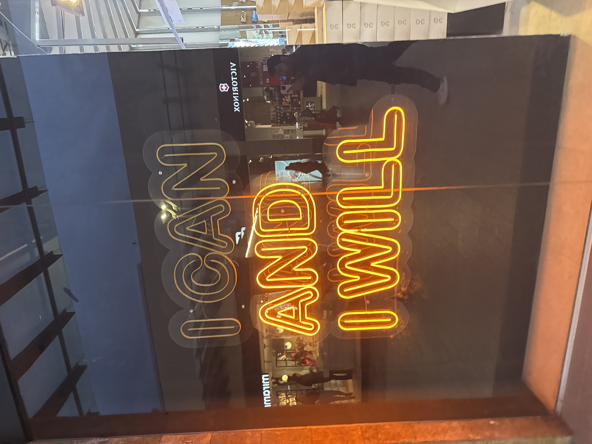 A daytime, indoor shot depicts a glass storefront in Puebla, Mexico, featuring a neon sign that reads "I CAN AND I WILL." The store displays the Victorinox logo above the neon sign. The foreground and background are captured in reflections within the glass. A person, possibly a customer, is seen inside the store, reflected in the glass. A row of boxes is positioned near the storefront's ceiling. The building's architecture includes a metal support structure visible on the left side of the storefront.
