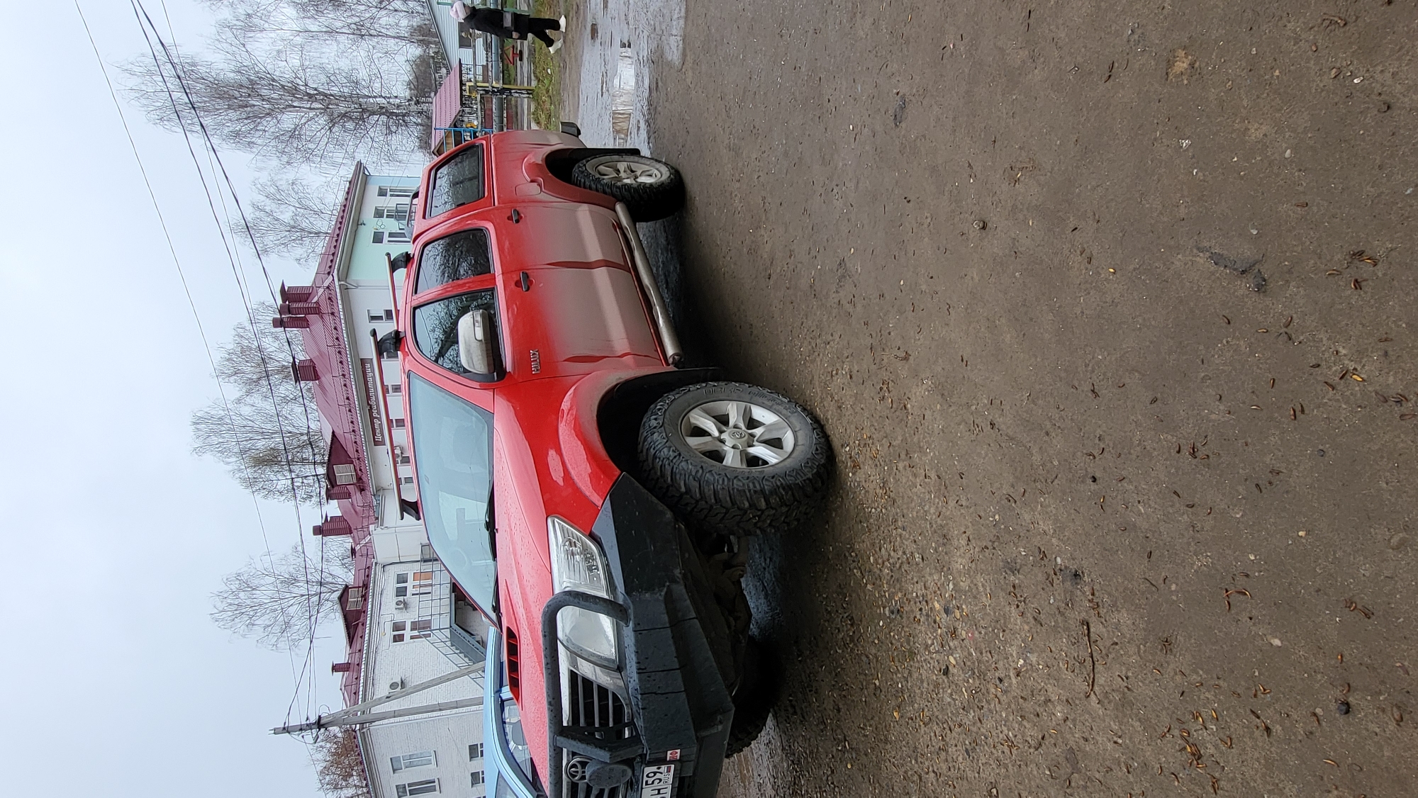 The image shows a red Toyota Hilux pickup truck, customized with a black bull bar and chunky off-road tires, parked on a wet, mottled surface. The truck is positioned on the right side of the frame, facing towards the left, and part of a blue car is visible in the bottom left corner. A partially obscured Russian license plate, displaying "H 59" and "RUS," is visible on the front of the red truck. The word "HILUX" is embossed on the truck's rear door.
The setting appears to be an urban or suburban area in Chaikovsky, Russia. The ground, a mix of asphalt and dirt, is damp or wet, with scattered debris and small leaves, suggesting recent rain or overcast conditions. In the background, under a cloudy, overcast sky, several buildings are visible. One prominent building features a red roof with multiple chimneys and a sign on its side with Cyrillic text that reads "Центр реабилитации" (Rehabilitation Center). Bare trees, indicative of late autumn, winter, or early spring, stand between the buildings and the cloudy sky. Power lines are visible stretching across the upper part of the scene. A lone person is walking in the distance on the wet ground, near what appears to be a small playground or park structure. The overall impression is of a quiet, damp daytime scene.