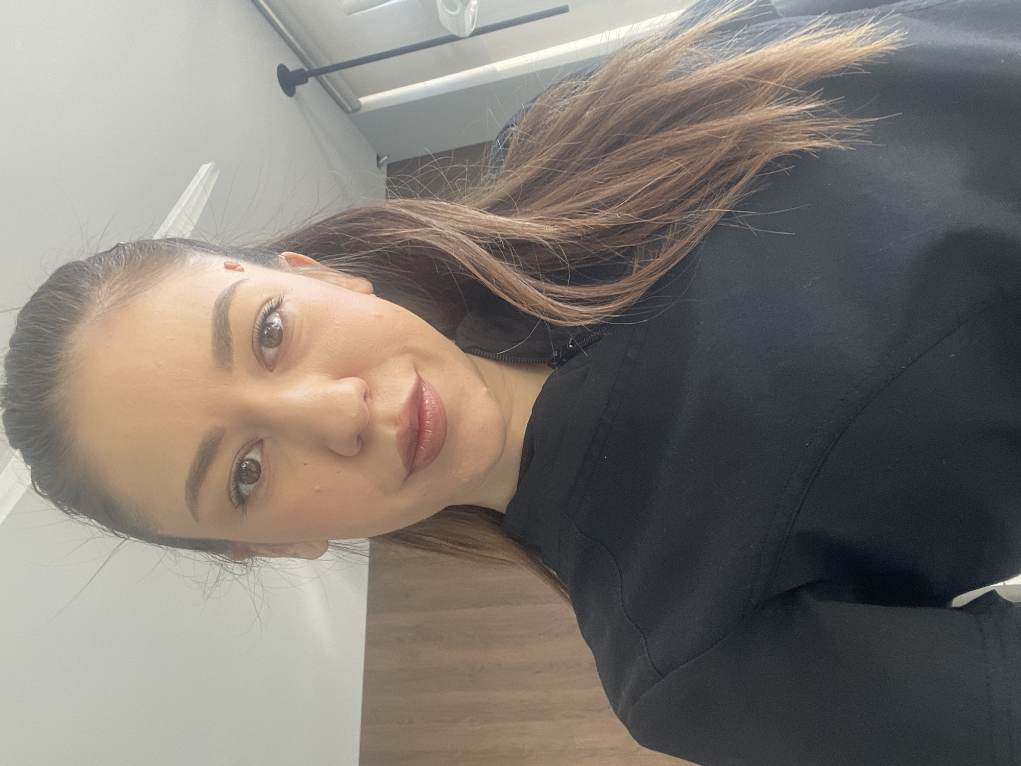 A medium shot depicts a woman with light brown hair and light skin wearing a black zippered top. She is facing the camera with a slight smile. The background consists of a light gray wall and wooden flooring. The image does not contain sufficient information to place the location in Sofia, Bulgaria.