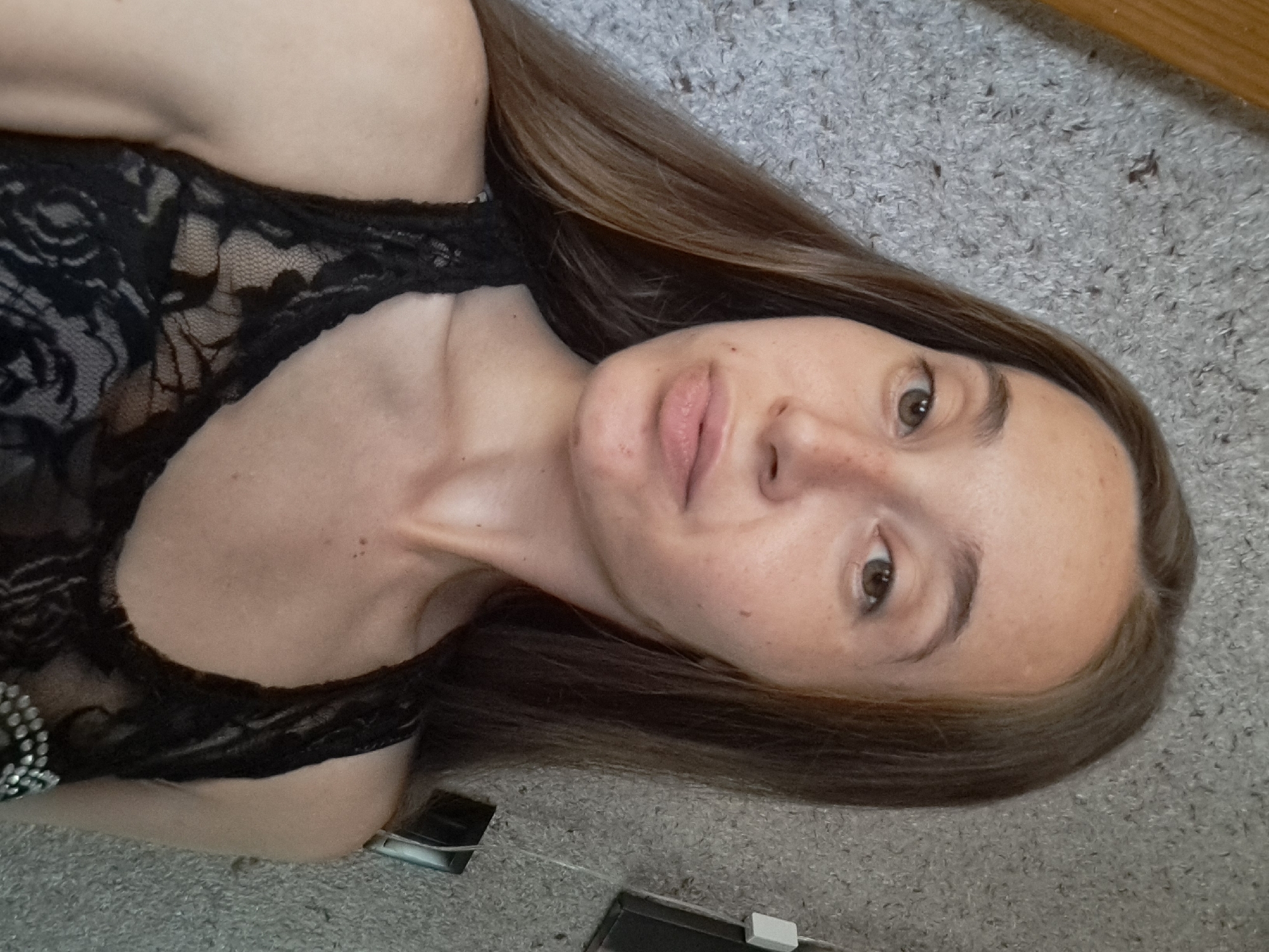 A young woman with long, brown hair and light skin is shown from the chest up, taking a selfie. She is wearing a black lace tank top. The background appears to be a textured grey wall. The image was captured in the city of Vypasne, Ukraine.