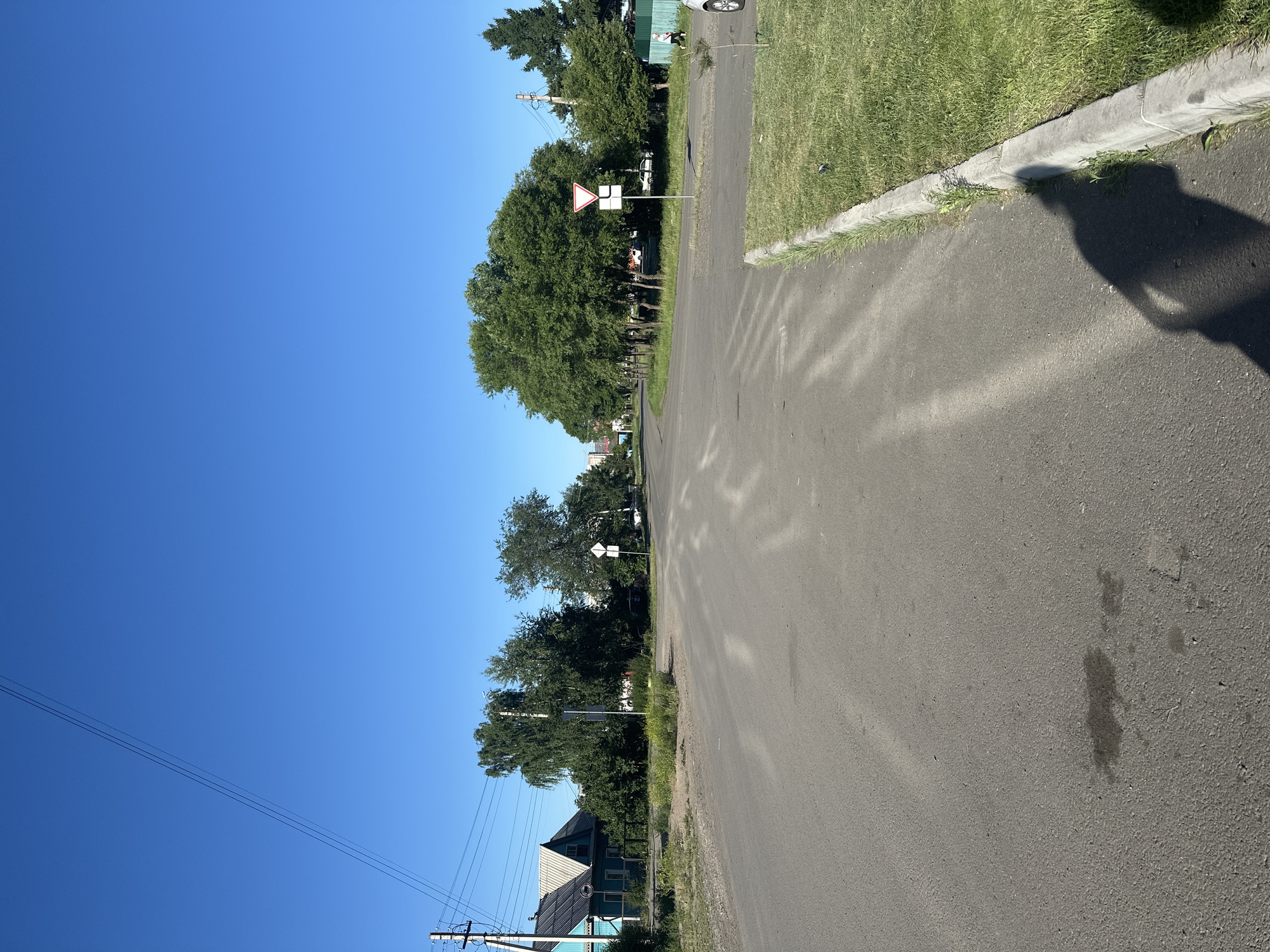 The image depicts a residential street scene in Kalinino, Russia, under a clear blue sky, suggesting a sunny day, likely in the morning or afternoon due to the shadows. The perspective is from ground level, looking down the asphalt road which curves gently to the right. On the left side of the road, a row of lush green trees lines the houses and properties. A prominent feature is a give way traffic sign with a white background and a red triangle. Further down the street, utility poles with power lines are visible, indicating a residential area. On the right side of the road, there is a patch of green grass with a concrete curb. A shadow, potentially cast by a person or an object, falls across the asphalt. The overall impression is a quiet, everyday scene in a rural or suburban setting.