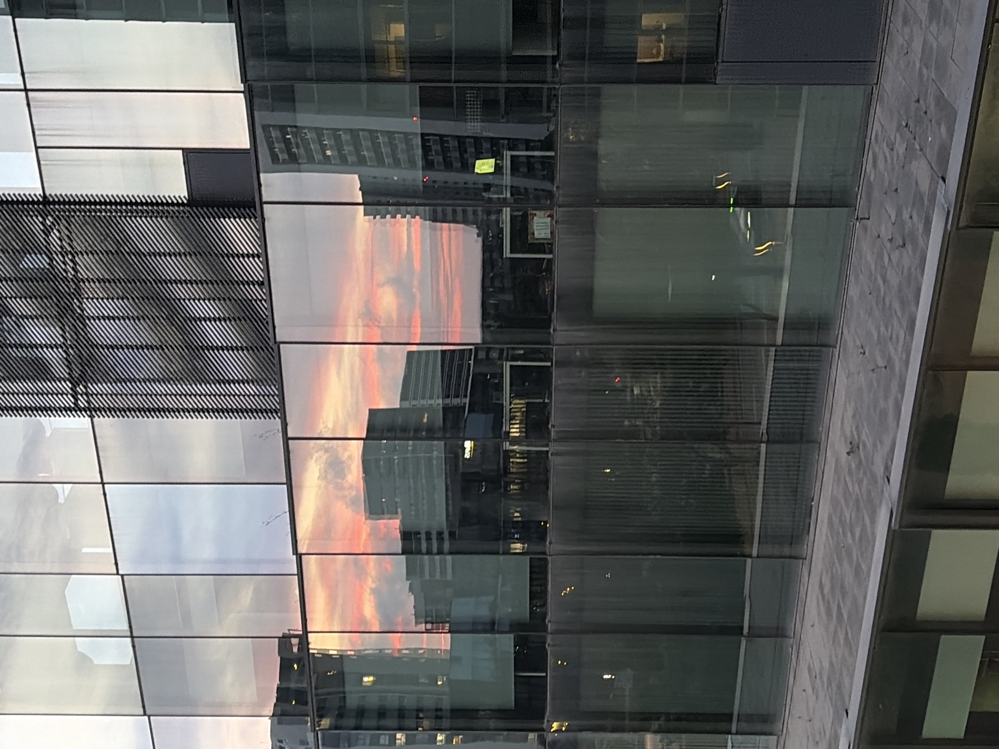 The image captures a close-up, upward-angled view of a modern glass-clad building, likely part of a cityscape in L'hospitalet De Llobregat, Spain. The primary subjects are the building's facade and the vivid reflections it casts.
The building's exterior is characterized by large, rectangular glass panels, some appearing clear and others with a darker tint or reflecting different elements. These panels are interspersed with dark, solid horizontal and vertical framing elements. A notable architectural detail includes a section on the left with a dark, textured, slatted, or grated surface.
The most striking feature is the reflection of a dramatic sunset sky in the glass panes. Vibrant streaks of orange, pink, and red clouds dominate several sections, contrasting with the cooler tones of the sky and other building reflections in adjacent panels. These reflections suggest it is either late afternoon or early evening. Other buildings, with their windows and structural lines, are also reflected, creating a complex interplay of real and mirrored forms within the urban environment. Some small, bright lights, possibly from within reflected buildings or the photographed structure, are visible as scattered points of light, enhancing the evening atmosphere.
No direct human activity is visible. The scene is static, focusing on the interplay of light, architecture, and reflection. The weather appears clear, allowing for the strong, colorful reflections.
Faint, illegible text appears in some reflected areas. One instance shows a small, pale yellow rectangular object, possibly a sticky note or sign, but its content is unreadable. Below it, another white rectangular sign with dark text is also too distant or blurry to be deciphered.