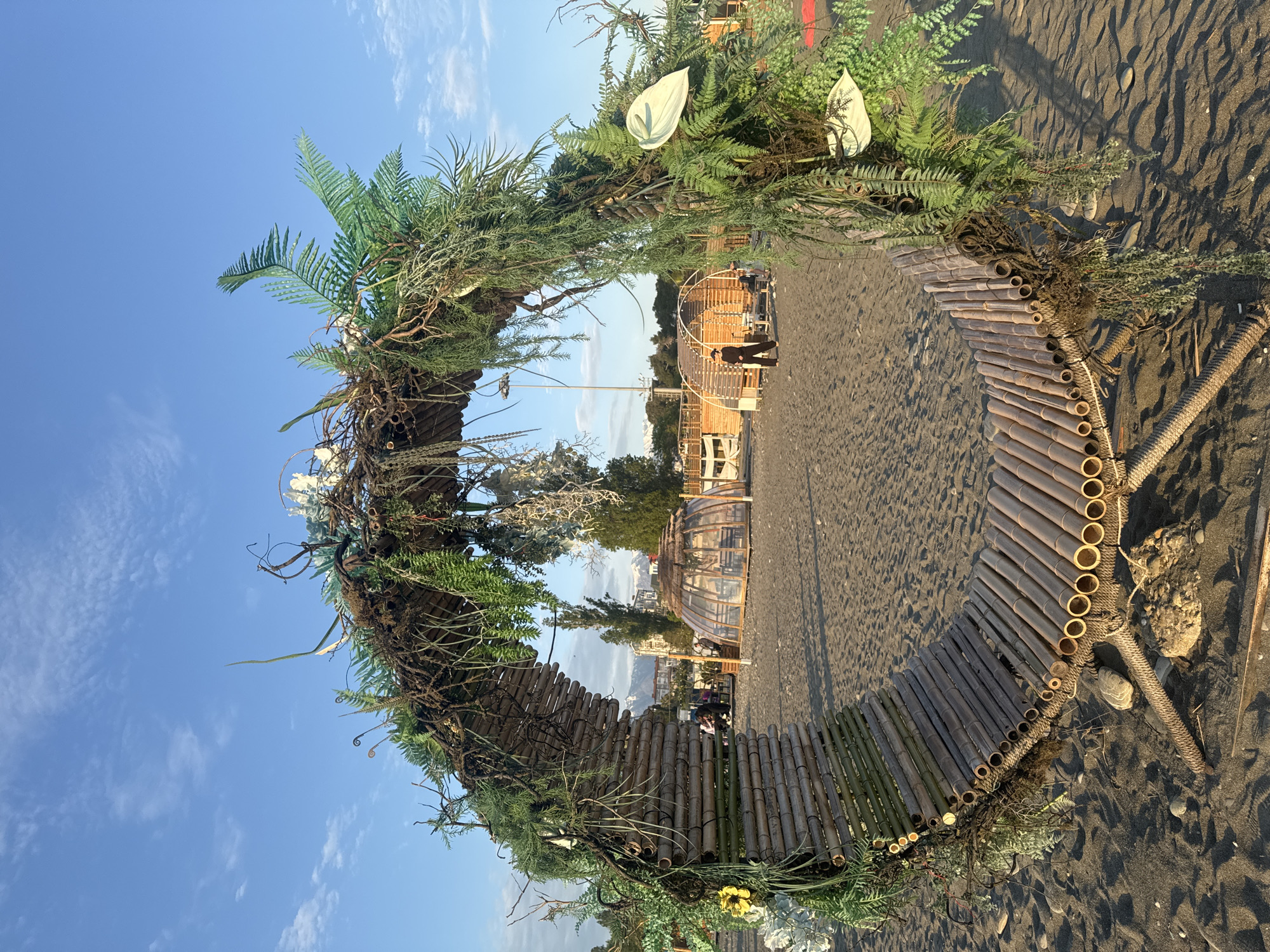 The image depicts a circular structure on a sandy beach in Sirius, Russia, under a clear, partly cloudy sky. The structure, made of bamboo and foliage, forms an archway or portal. The base of the structure is constructed from bamboo poles laid horizontally and bound together, creating a circular path or platform on the sand. Green foliage, including ferns and other plants, is interwoven into the structure, giving it a natural, organic appearance.
Through the center of the circular structure, a view opens to a scene including buildings and possibly other temporary structures. There are also at least two people in the background. The sand is dark, possibly volcanic. The lighting suggests it is either early morning or late afternoon.