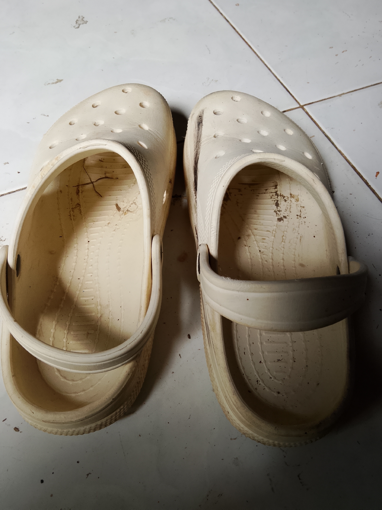 Two light-colored clogs, showing signs of wear and dirt, are placed on a tiled floor in Merbopuntong, Indonesia. The clogs are made of a rubber-like material and have a classic clog design with ventilation holes on the top. The left clog is positioned slightly ahead of the right clog, and both are facing upwards. The floor is made of light-colored square tiles, with visible grout lines. There are small dark specks and some debris scattered on the floor and inside the clogs, indicating a lived-in environment. The lighting in the scene is somewhat dim, casting shadows around the clogs.