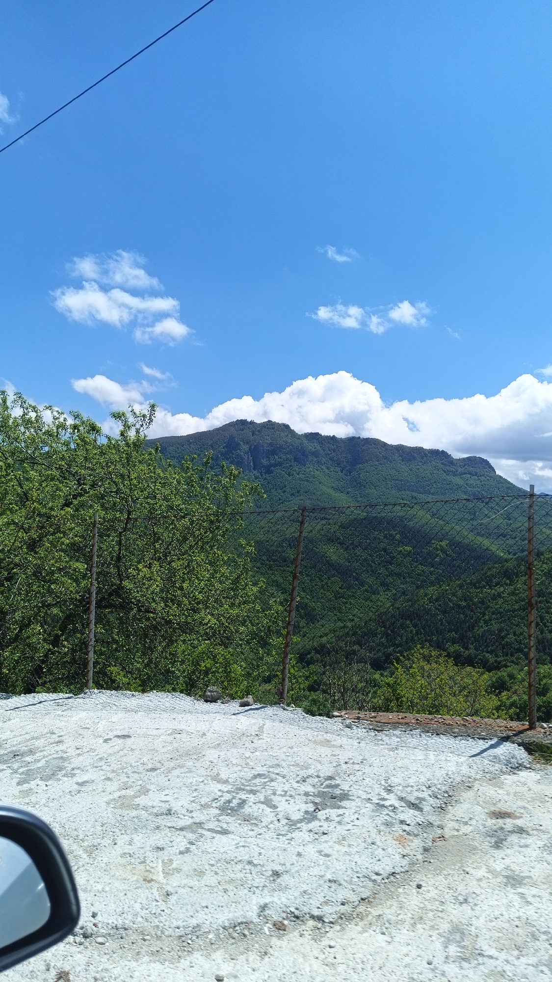A sunny day in Oreshets, Bulgaria, featuring a view of a lush, green mountain partially obscured by trees and a wire fence. Above the mountain, a bright blue sky is dotted with puffy white clouds. In the foreground, there's a weathered concrete surface. The side mirror of a vehicle is visible in the bottom left corner, suggesting the image was taken from inside a car. An electrical wire stretches across the top of the frame.