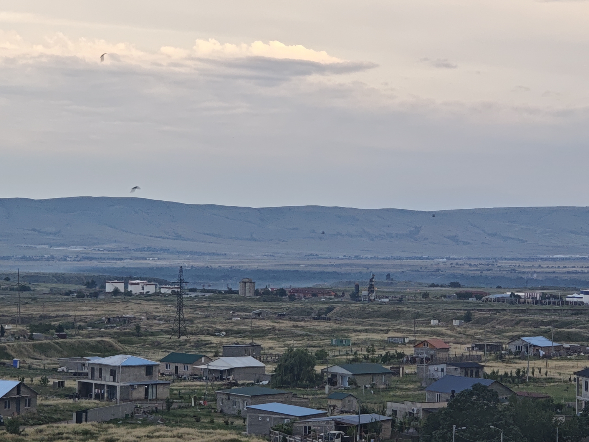 This media file is a photograph of a rural or suburban landscape with a village nestled at the foot of a mountain range. The scene is bathed in the soft, diffused light of either dawn or dusk, indicated by the muted colors and the overcast sky. The weather appears to be calm, with no strong winds or precipitation visible.
The foreground is dominated by a collection of houses, varying in architectural style and size, suggesting a developing community. Some houses are simple, single-story structures, while others are larger, multi-story buildings with blue and gray roofs. The surrounding land is a mix of dry grass and sparse vegetation, with a few trees scattered throughout. Utility poles and power lines are visible, indicating the presence of infrastructure.
In the middle ground, there are larger industrial or agricultural buildings, including several white tanks with red tops, a tall transmission tower, and other structures that appear to be part of a complex. This juxtaposition of residential and industrial elements suggests a mixed-use area.
The background features a long, rolling mountain range that stretches across the horizon. The mountains are a muted blue-gray, softened by the distance and the atmospheric conditions. A few distant buildings or structures can be seen on the plains between the village and the mountains.
Two birds are captured in flight against the cloudy sky, adding a touch of dynamism to the serene landscape. The overall mood is one of quietude and rural living, with a hint of industrial presence in the distance. There is no visible text in the image. The location context provided is Tbilisi, Georgia.