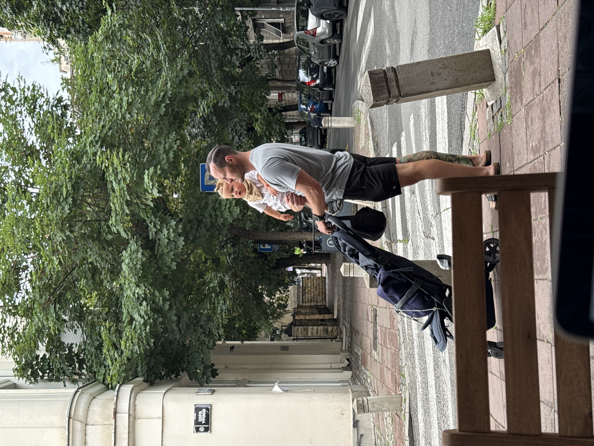 A man is standing on a sidewalk in Belgrade, Serbia, holding a baby. The man is wearing a grey t-shirt and black shorts. The baby is wearing a white shirt and a tan scarf. The man is holding a stroller with a blue bag in it. The sidewalk is paved with red bricks. There is a street with cars parked on the side. There are trees and buildings in the background. A street sign is visible on the left side of the image, which reads "ЦАРА УРОША".